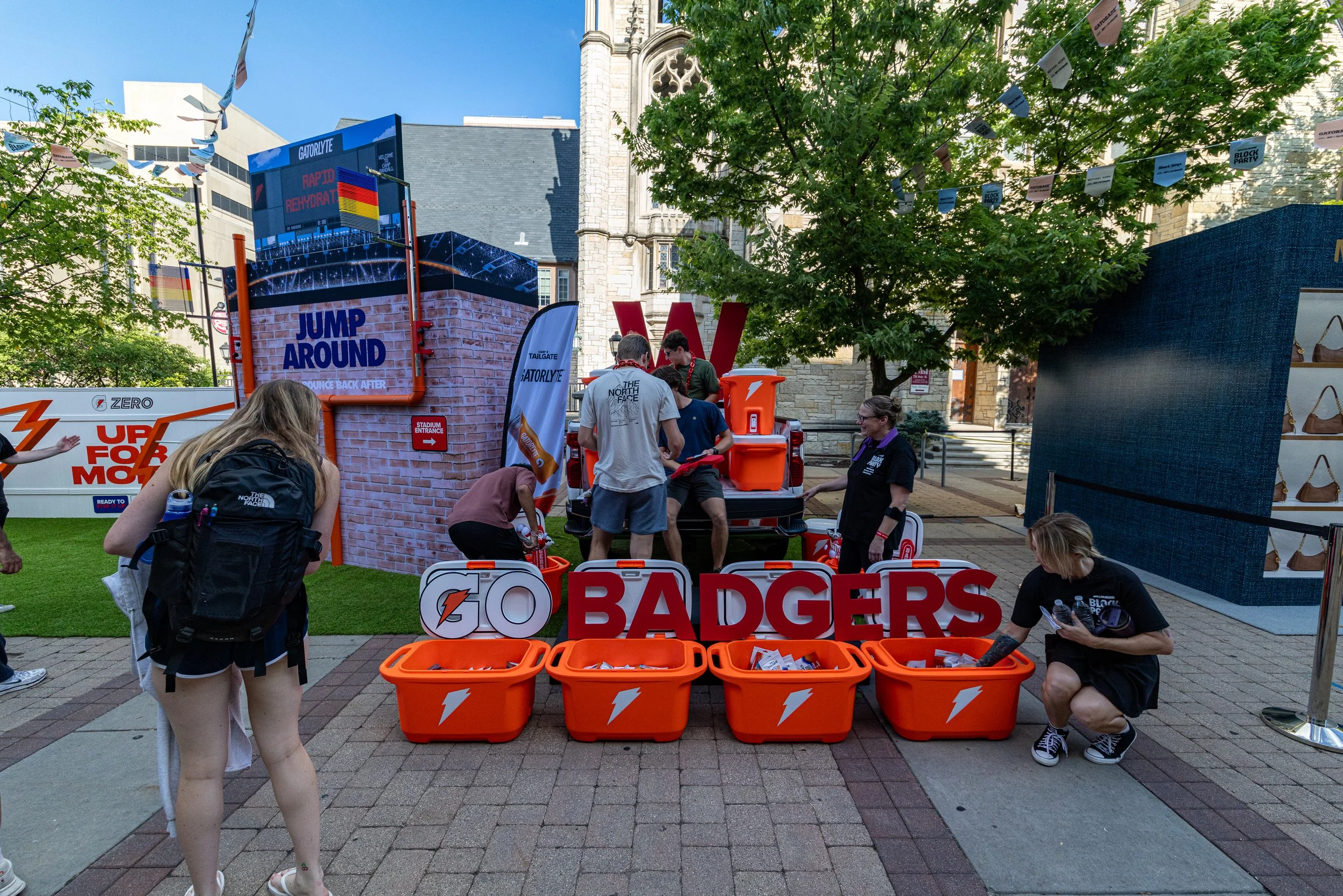 People gathering at an outdoor event with orange bins labeled 'Badgers' and 'Gatorade' boxes, a large digital screen displaying sports-related graphics, and several banners and signs.