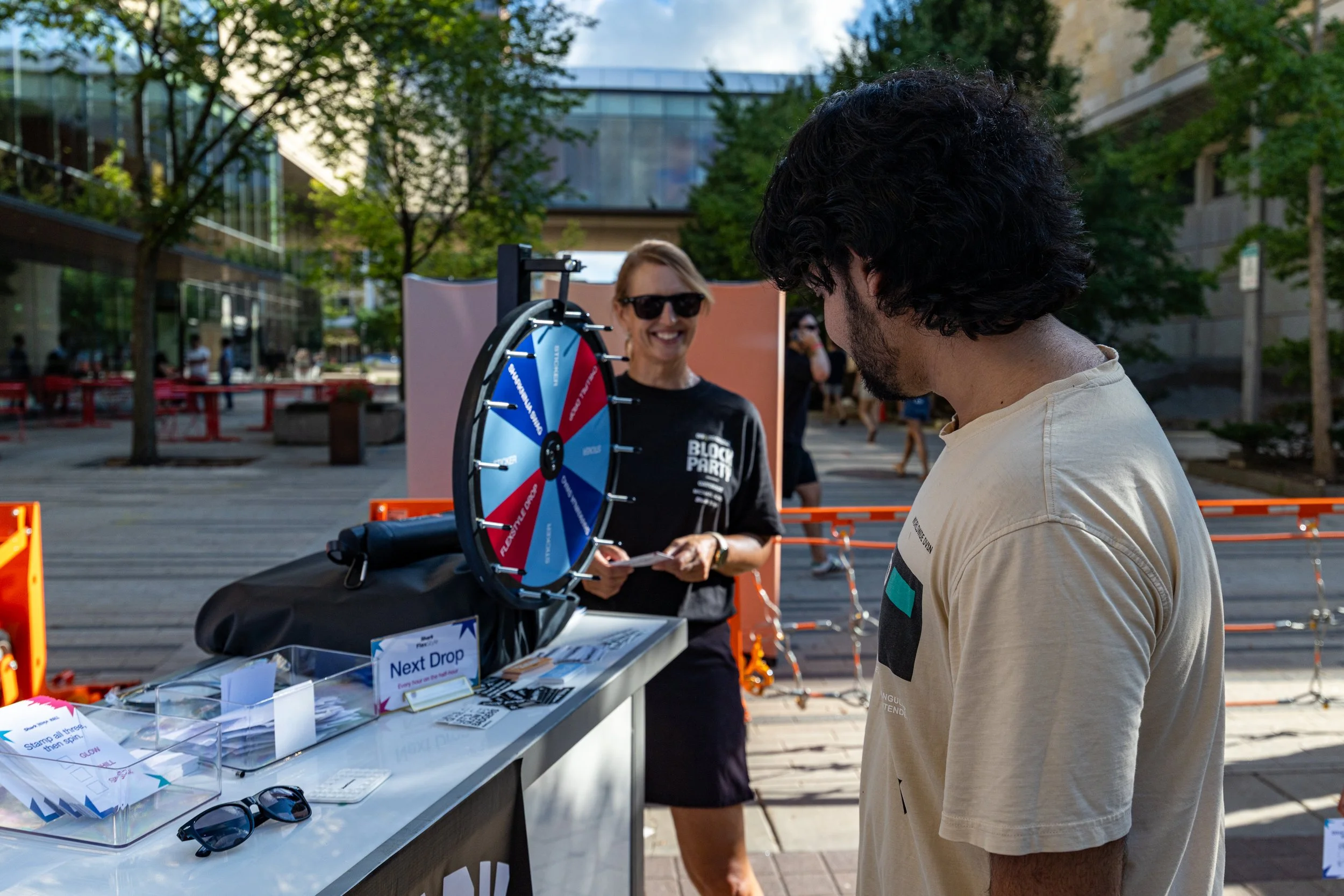 A woman smiling behind a spinning wheel game at an outdoor event, with a man standing in front of her. The woman has blonde hair and wears sunglasses, and the man has dark, curly hair. There is a table with papers, sunglasses, and a sign that says "N