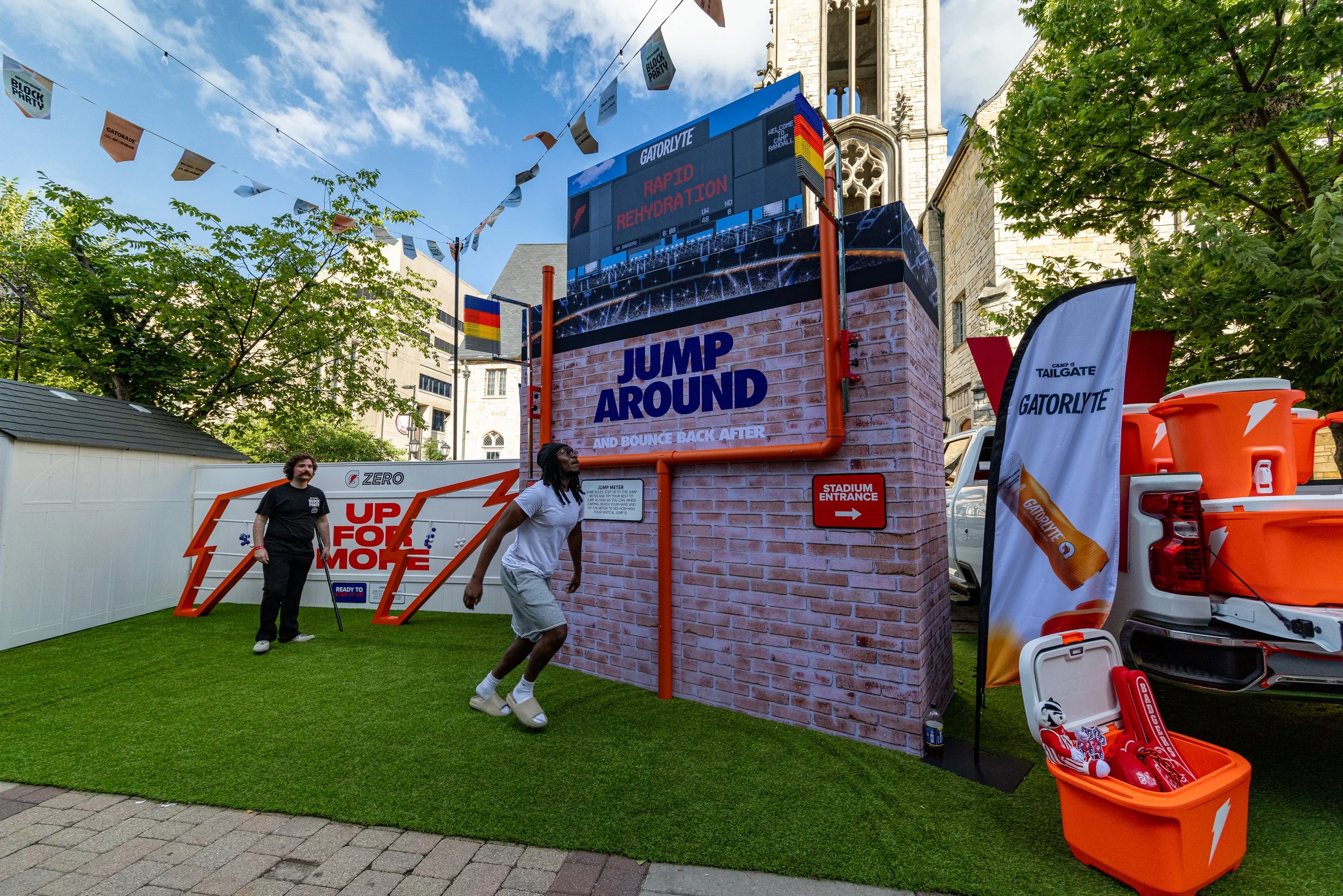 People participating in an outdoor event with Gatorade branding. A large electronic sign displays 'Rapid Rehydration' and a scoreboard. There are flags, a tent, and equipment related to sports hydration and recovery, with a church in the background a