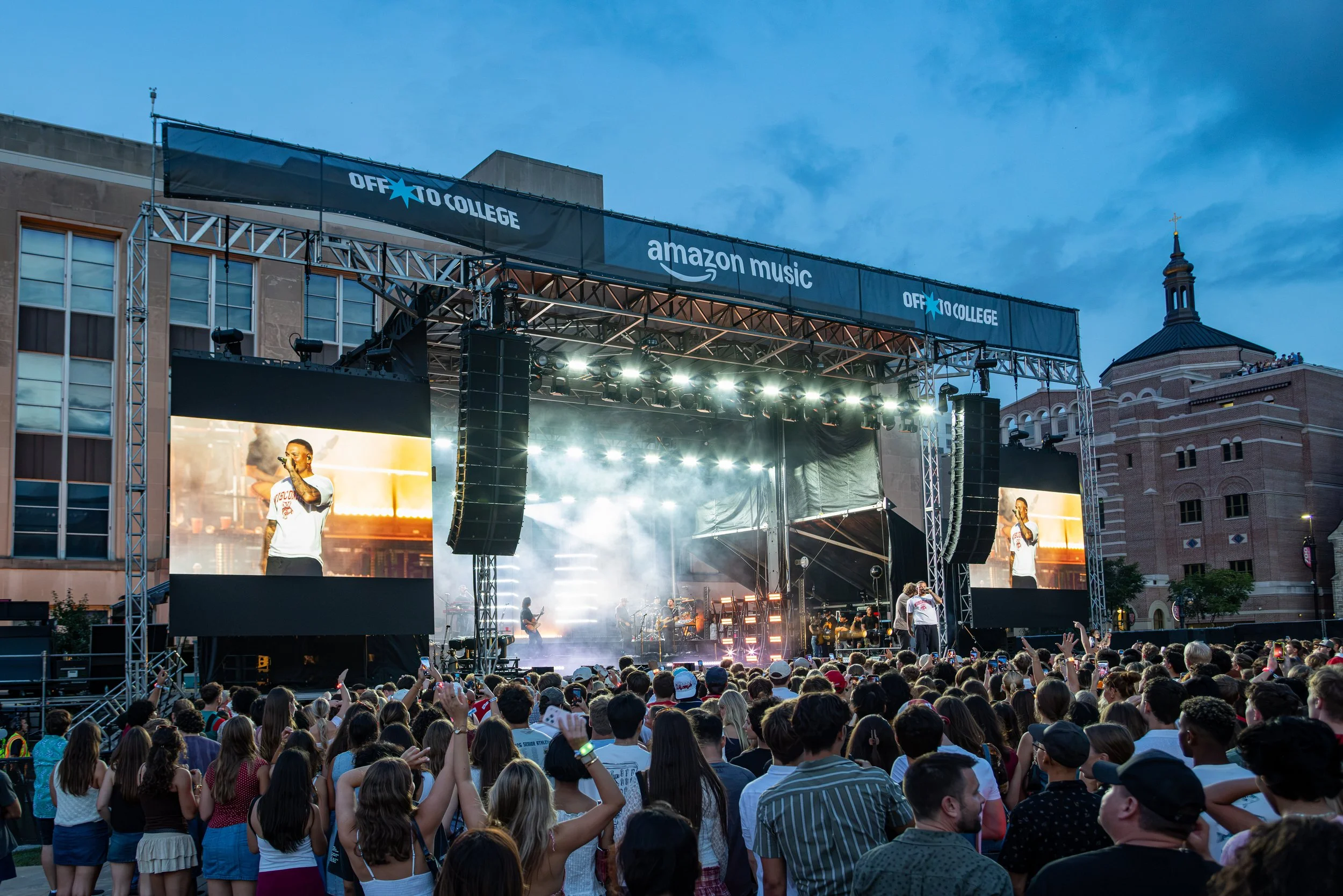 Outdoor concert with a large crowd watching a stage performance at sunset. The stage has screens displaying performers and banners with 