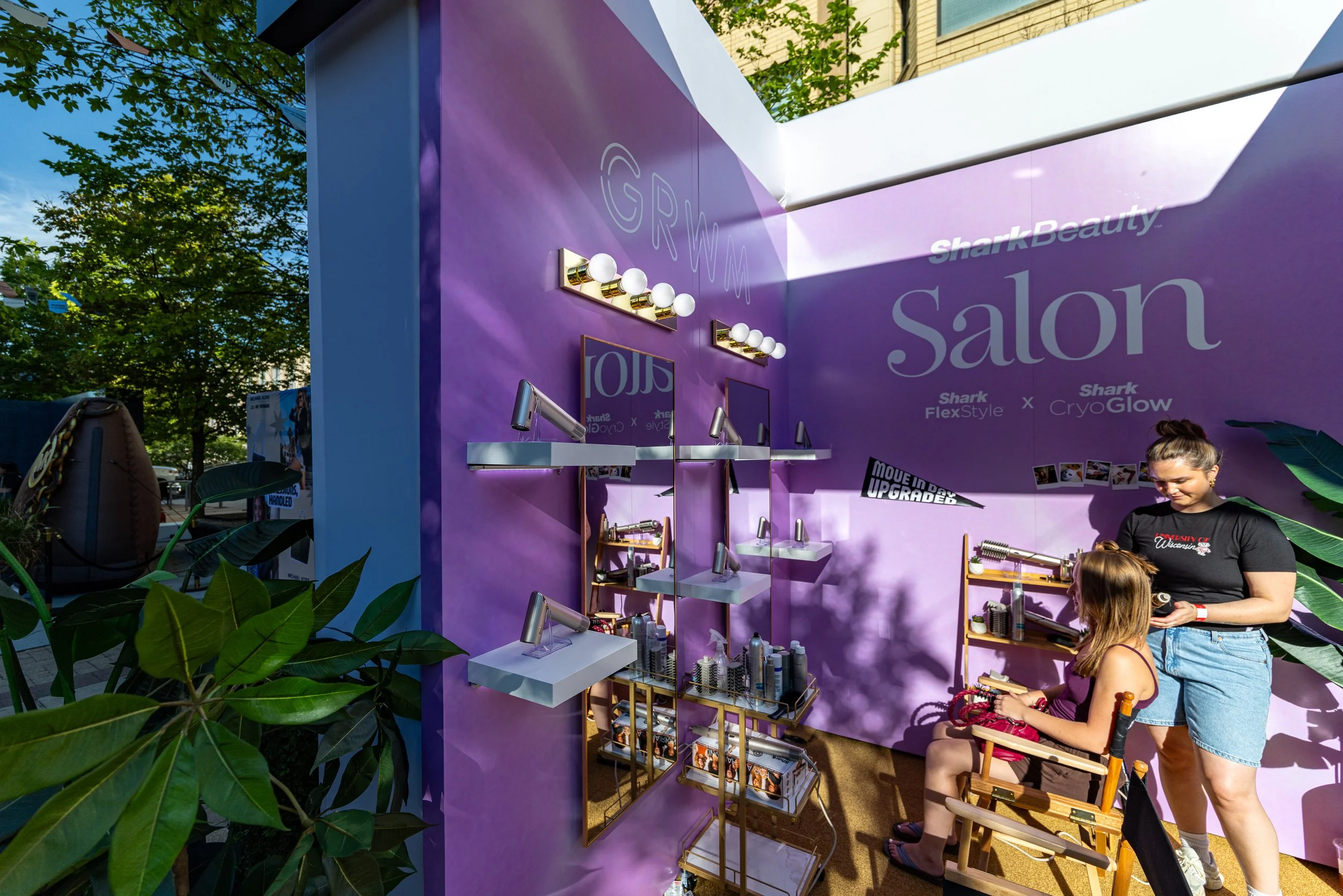 At an outdoor beauty booth, two women are having a makeup session. One woman is seated, and another woman is standing, applying makeup. The booth has a purple wall with the words 'Shark Beauty' and 'Salon' and various beauty products displayed on she