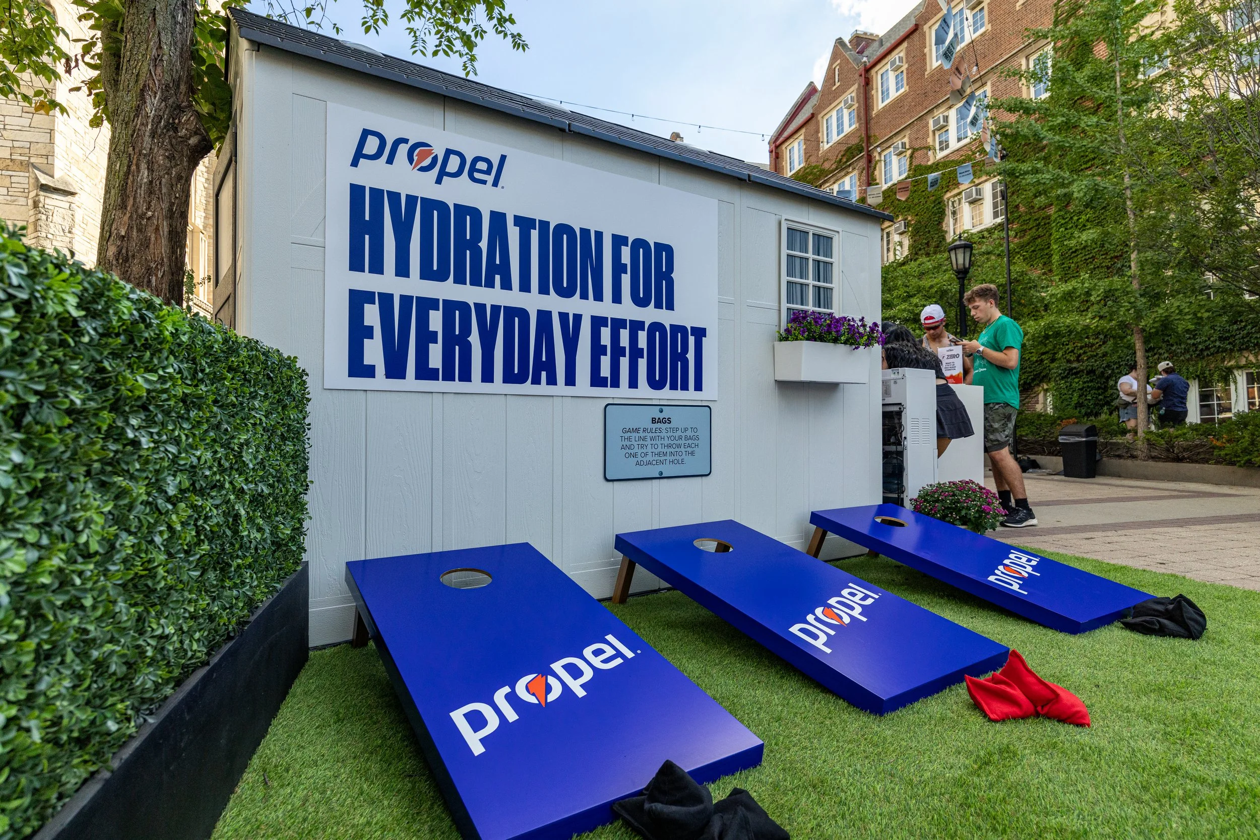Outdoor hydration station set up with three blue Coroplast boards with 'Propel' logo, a white structure with a sign reading 'Hydration for Every Day Effort,' surrounded by green bushes and trees, and people in casual attire around the station, with b