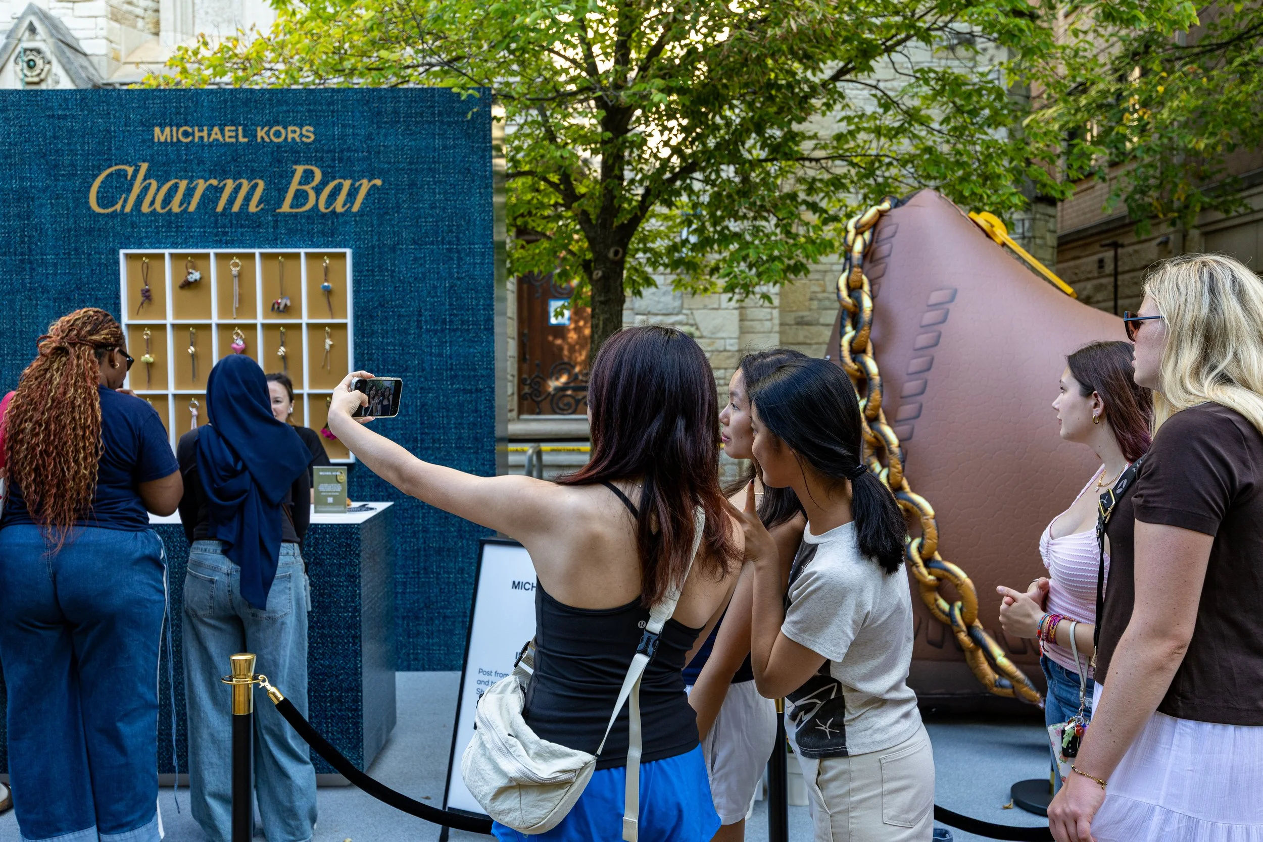 Group of people standing in line at a Michael Kors Charm Bar event, with a large decorative shoe in the background and a display of charm jewelry.