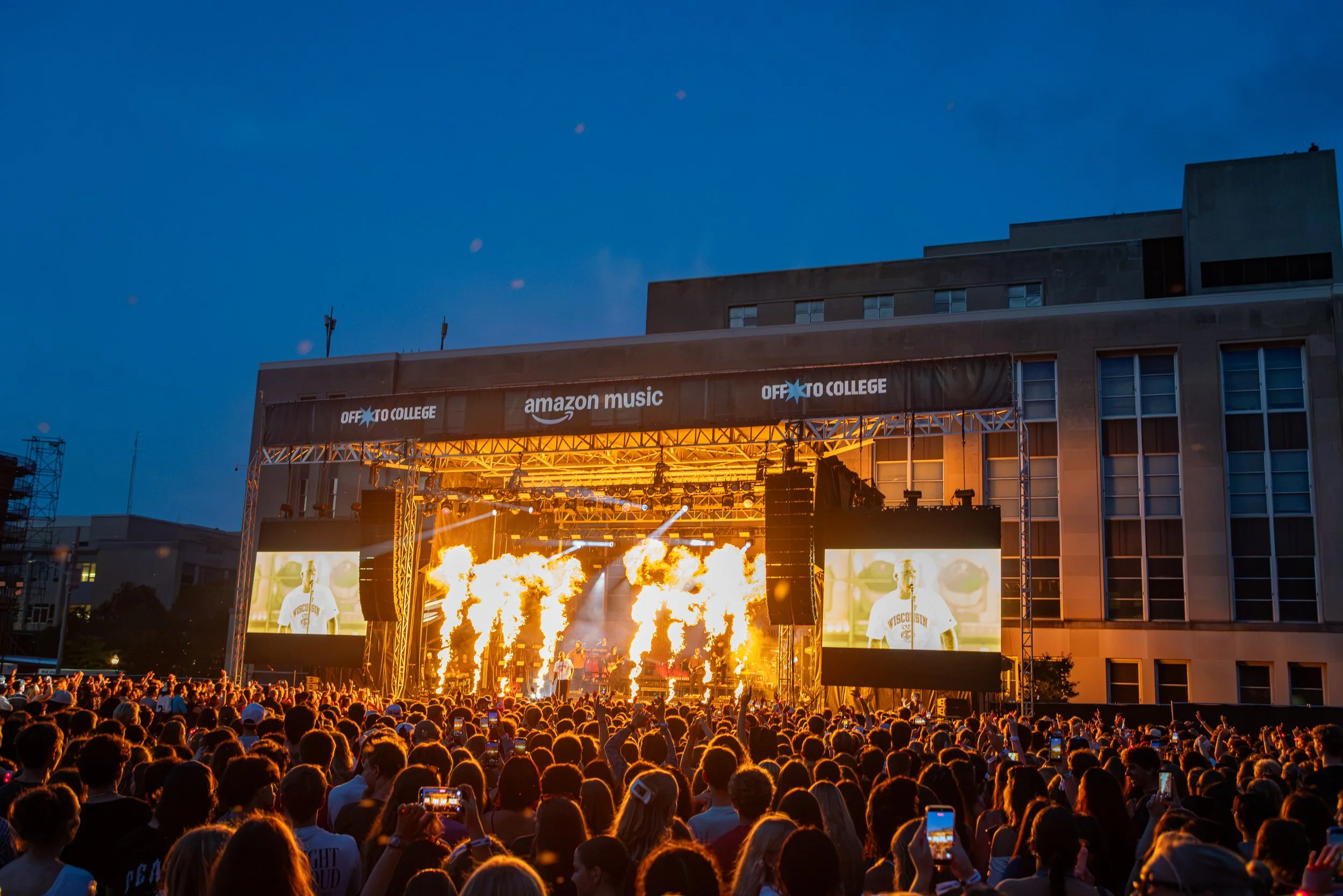 A large crowd watching a concert outdoors at dusk with a stage featuring pyrotechnics and large screens, and signs reading 'OFF TO COLLEGE' and 'amazon music'.