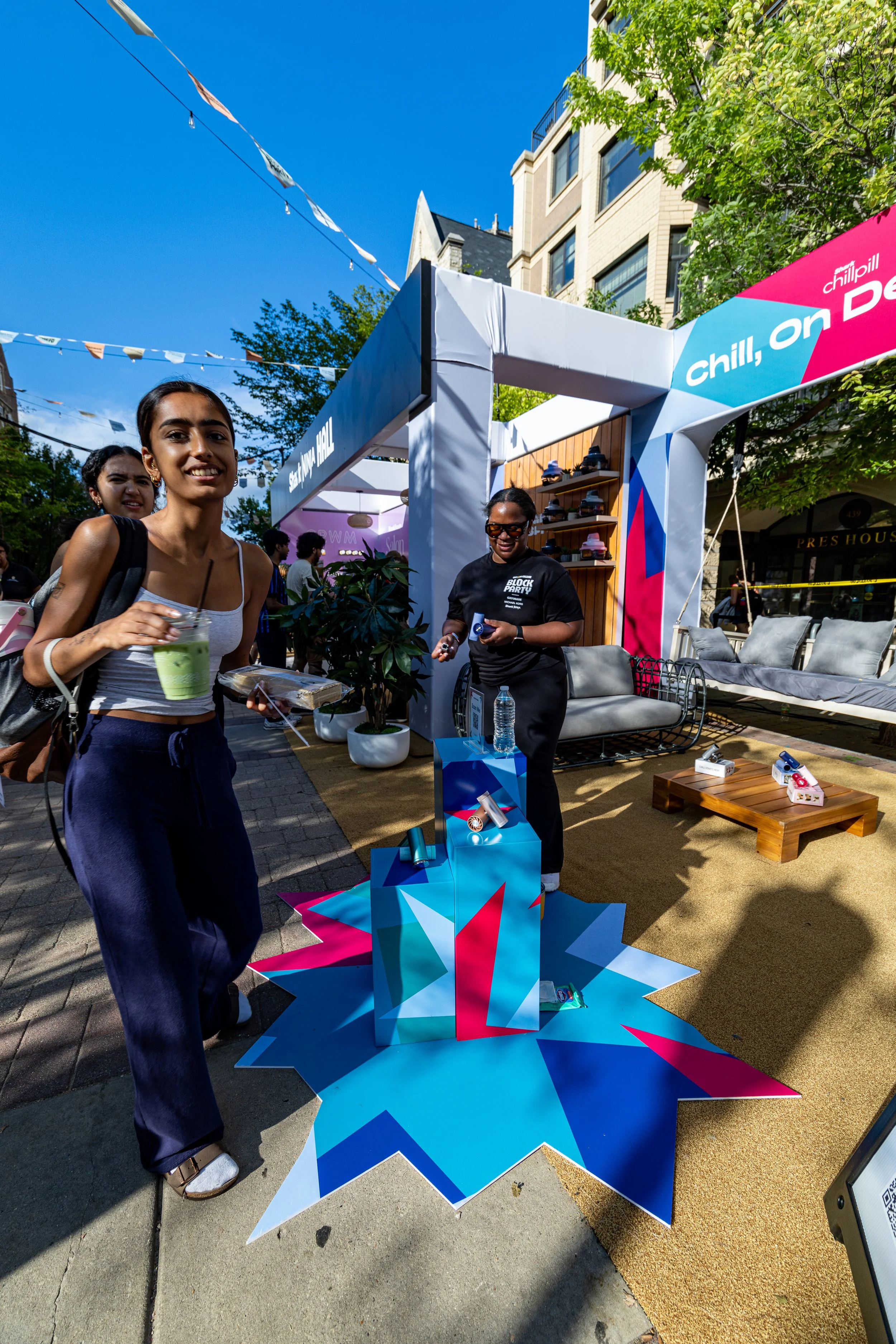 Street scene at an outdoor event with a colorful geometric star-shaped display on the sidewalk. Two women are in the foreground, one smiling and holding a green drink, and the other smiling and standing behind a booth. The booth has banners with the 