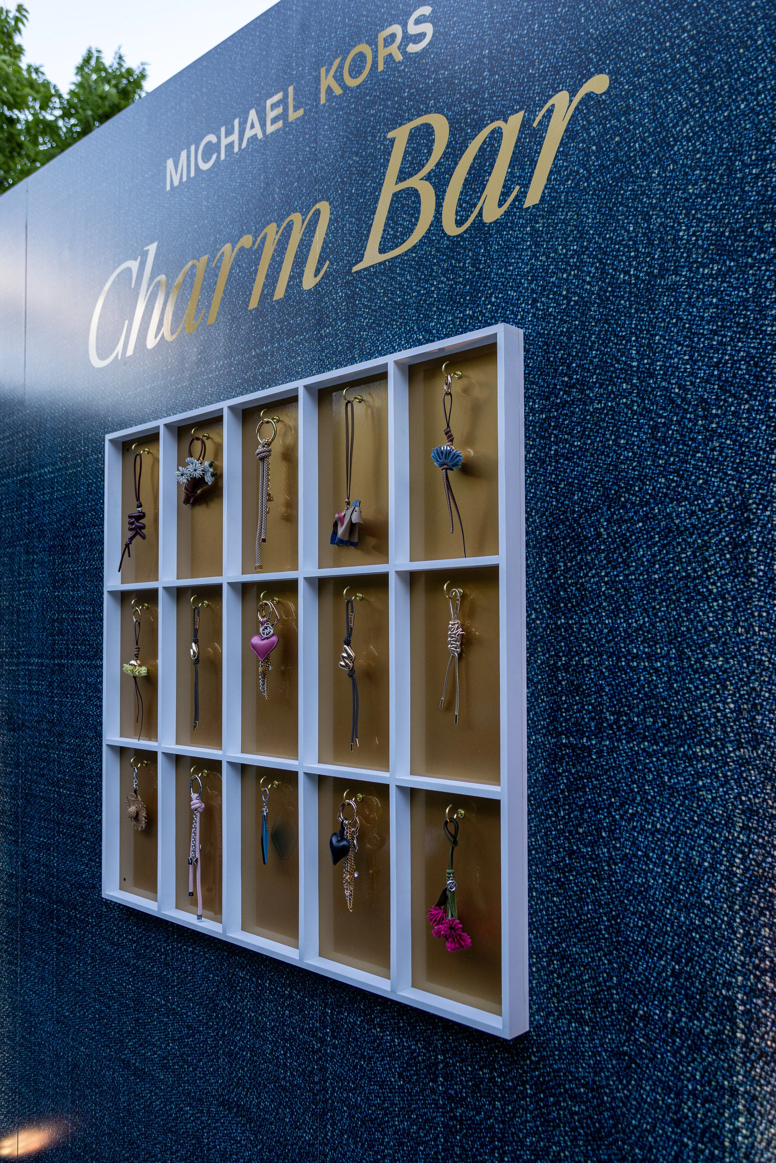Display board with various jewelry items, including necklaces and earrings, against a textured blue background. The board features the name 'Michael Kors' and the phrase 'Charmed By!'