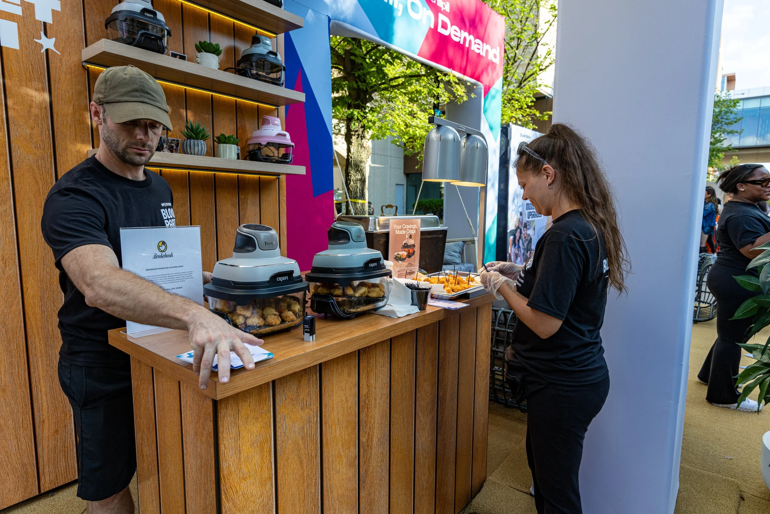 A man and a woman serve food at a booth in an outdoor event. The man wears a khaki cap and black t-shirt, and the woman has glasses and is wearing black as well. The booth features a wooden wall, shelves with small appliances, potted plants, and a si