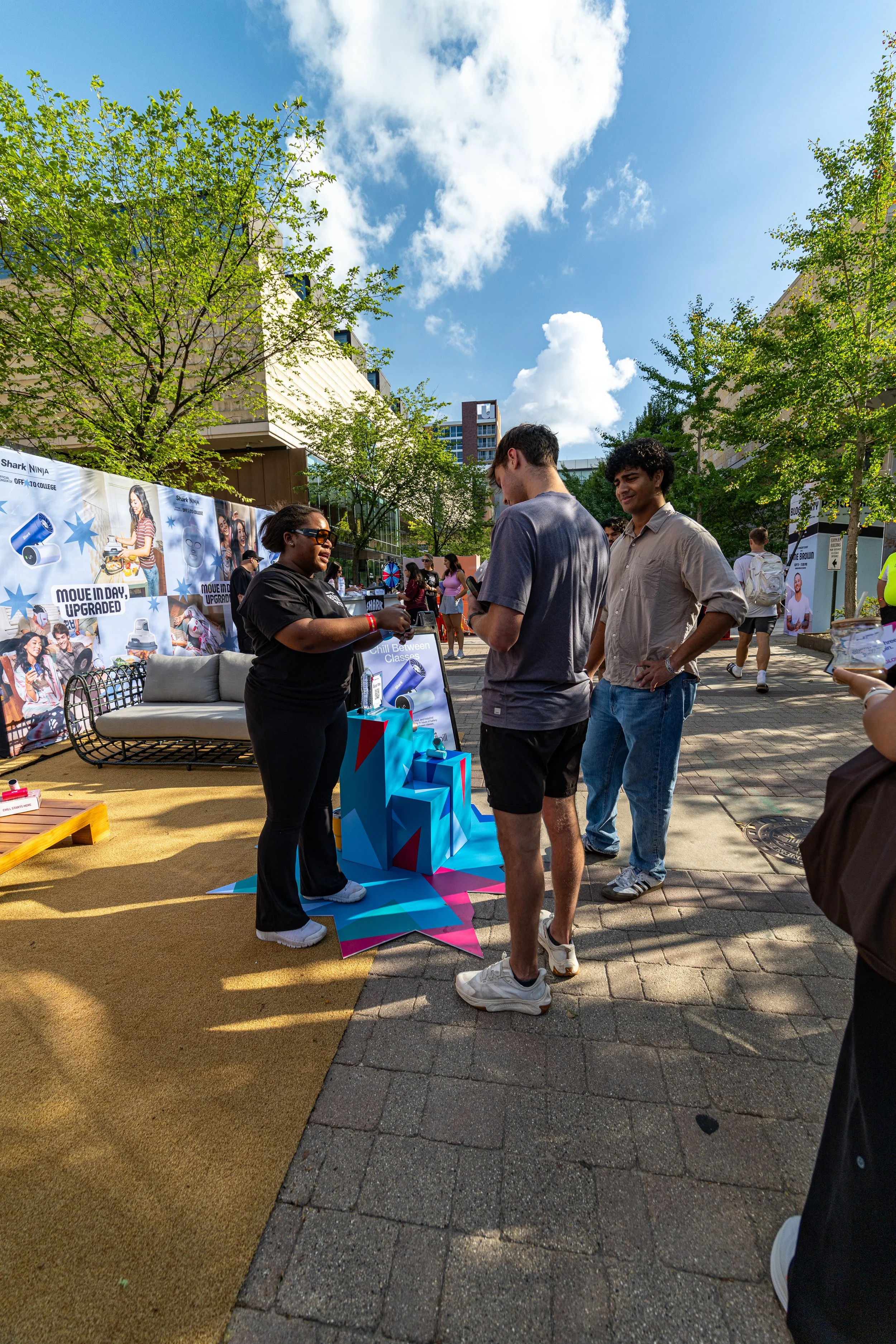 People gathered outdoors at a booth with promotional materials, trees, and a city skyline under a partly cloudy sky.