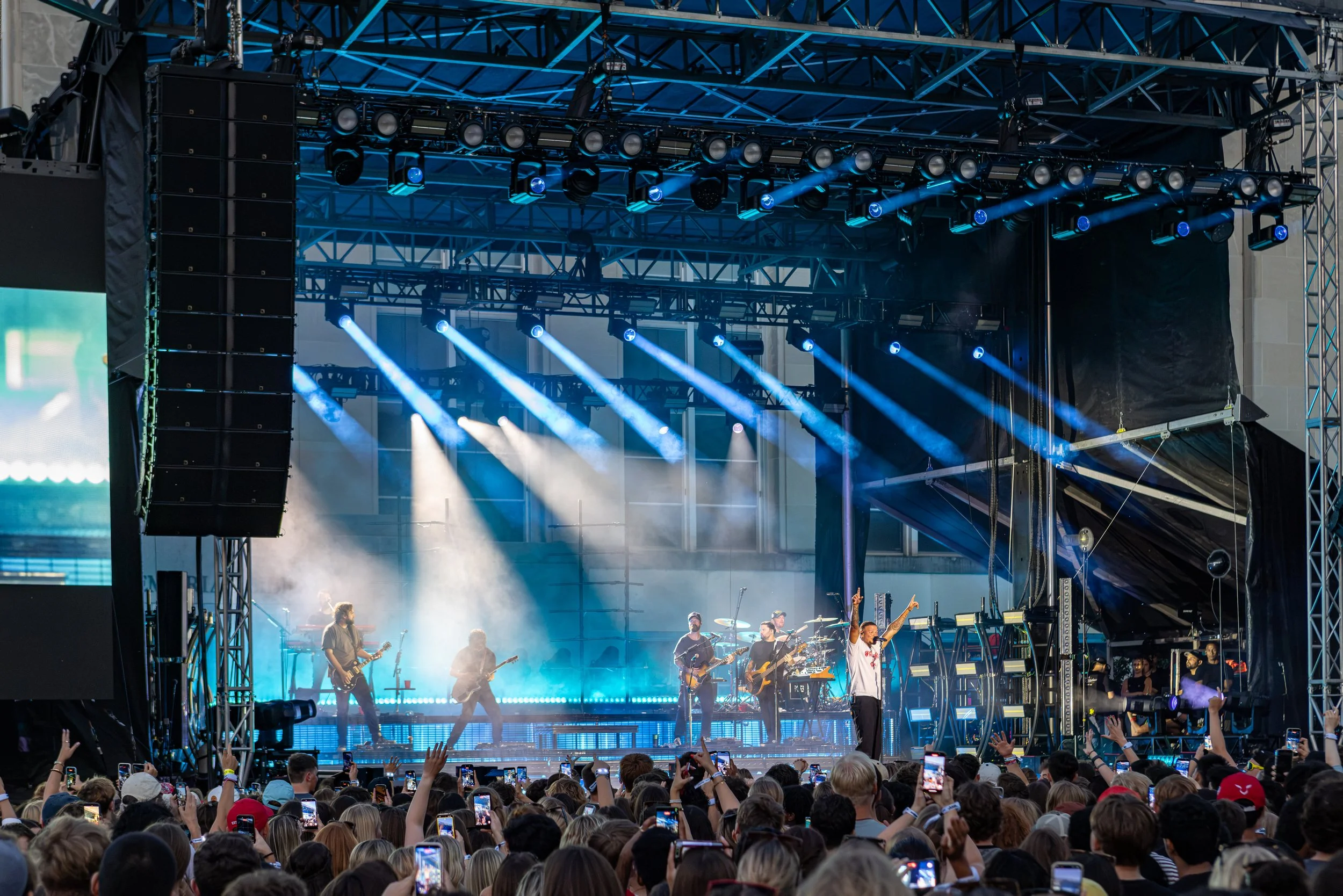 A large outdoor concert stage with bright blue and white lighting, a band performing, and a crowd of people taking photos and watching.