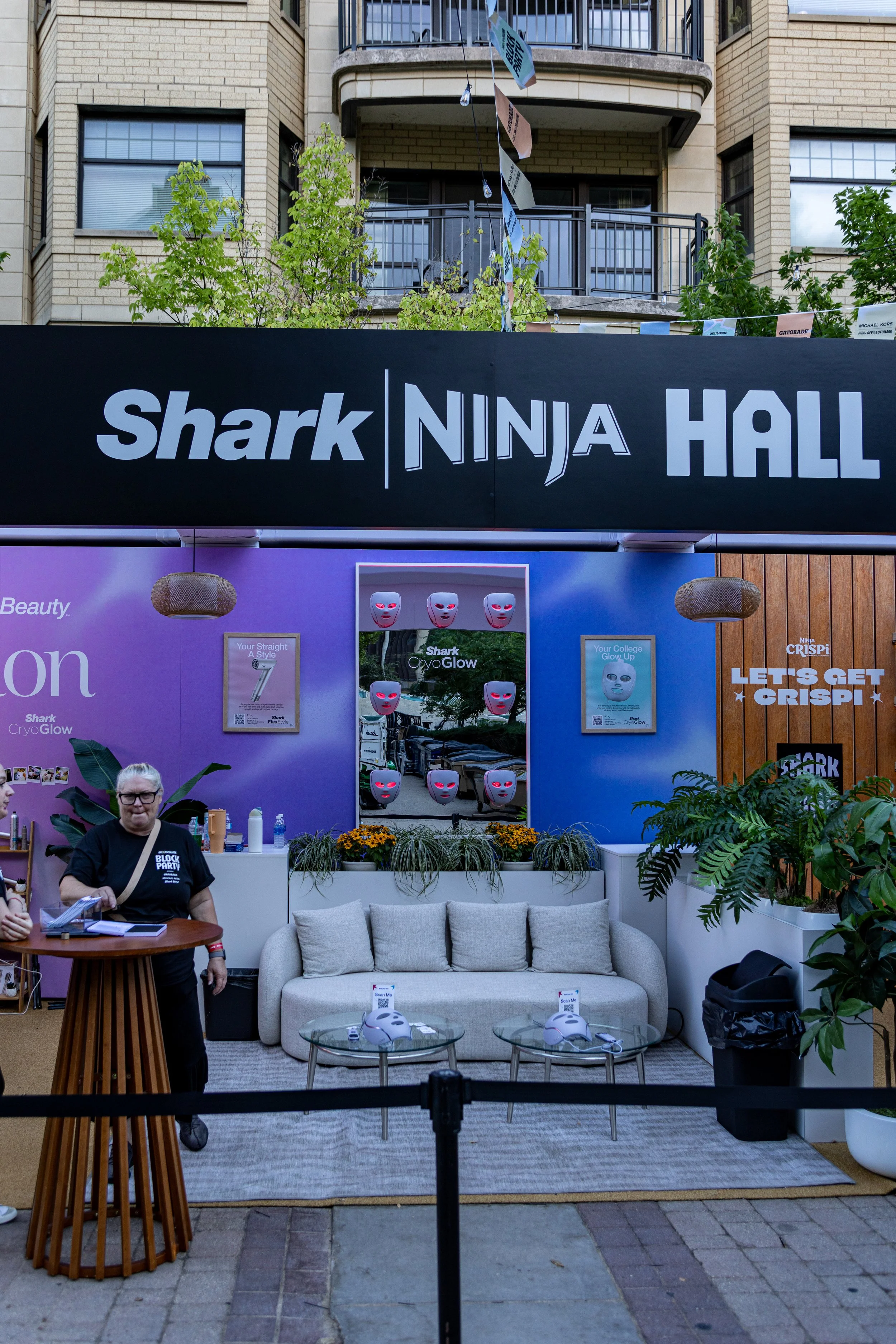 A display booth with a black sign that reads 'Shark NINJA HALL' over a seating area with a beige sofa, glass-topped coffee table, and plants. There are posters and masks related to Shark and Ninja products on the purple and orange walls, and two whit