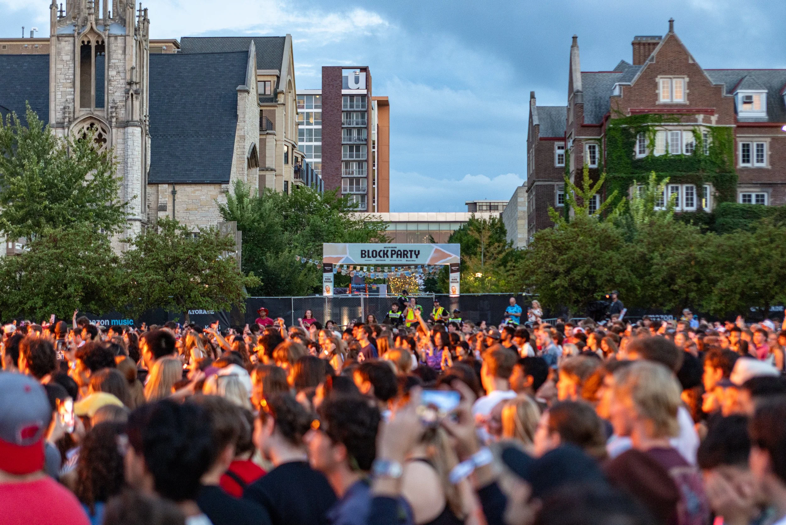 Crowd gathered outdoors for an event, with stage setup and people watching, city buildings and cloudy sky in the background.