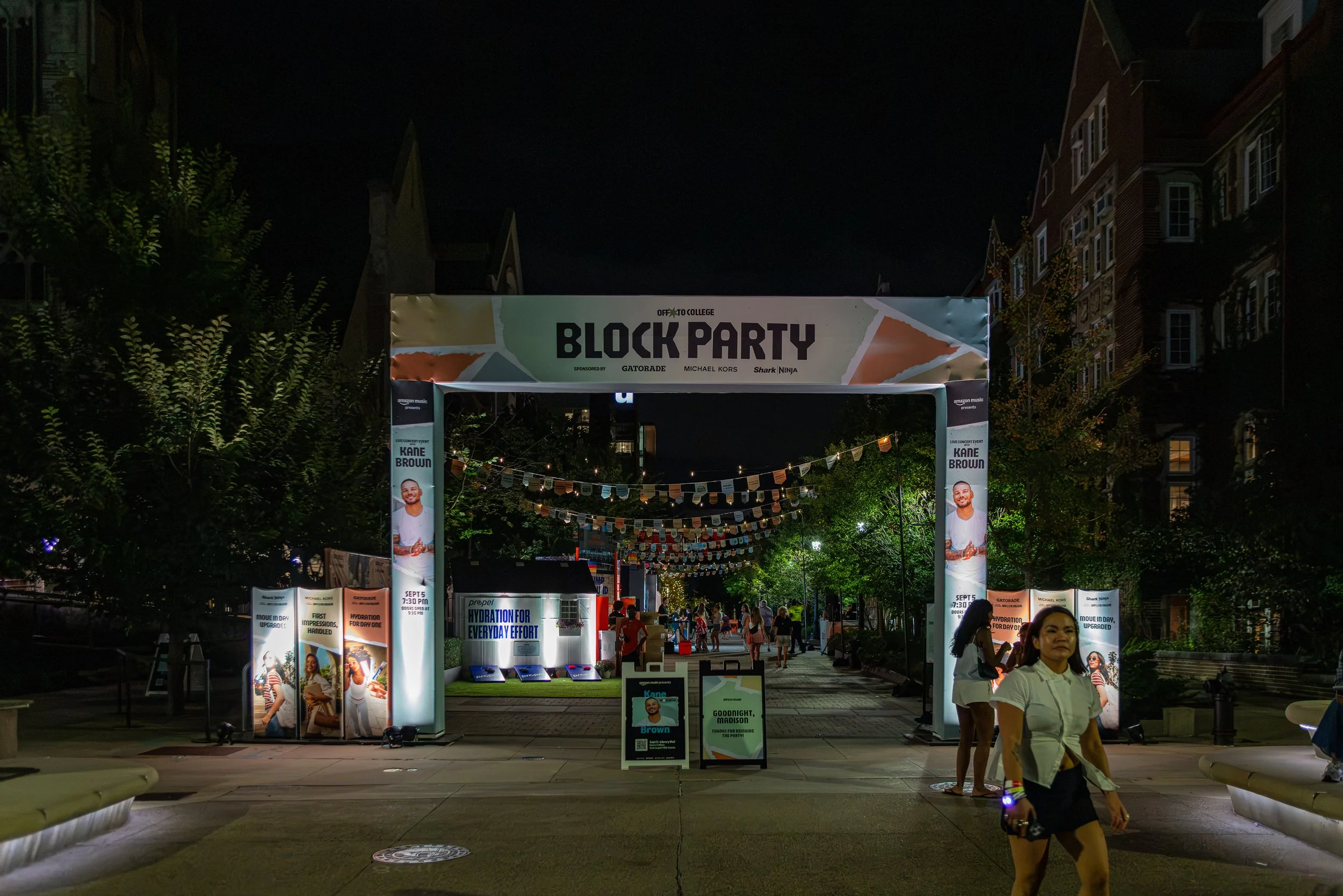 Nighttime scene of an outdoor event called 'Block Party' with a large entrance sign featuring a colorful geometric design. The event is sponsored by brands like Gatorade, Michael Kors, and Shark Ninja, and has images of people, including a man named 