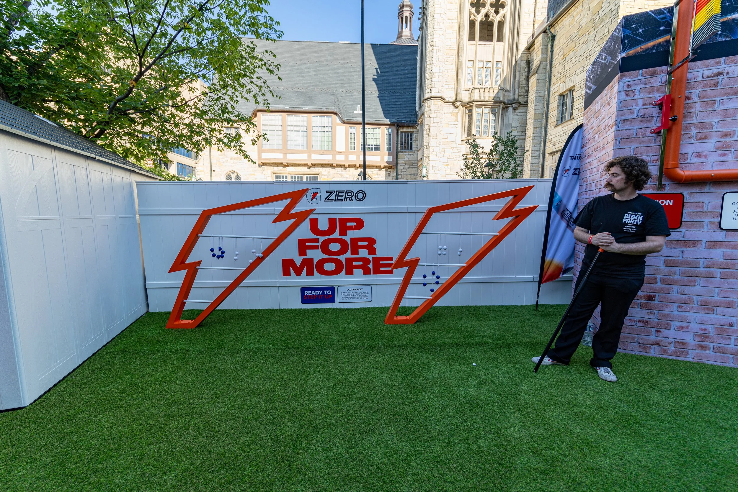Man standing on artificial grass near a shot basketball goal with a sign that reads 'UP FOR MORE' in front of a white fence. A brick wall and a historical building with large windows are in the background.