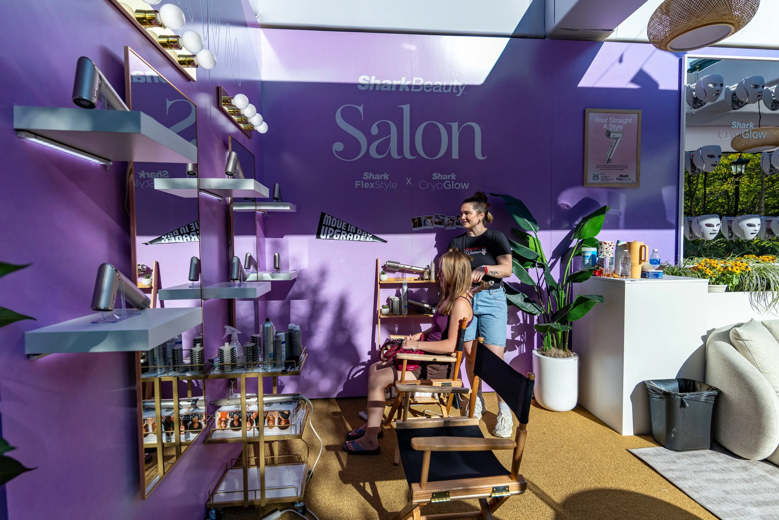 A beauty salon setup featuring purple walls with large text advertising 'Shark Beauty' and 'Salon', and smaller text about 'Shark FlexStyle' and 'Shark CryoGlow'. There are multiple mirrors with white spherical light bulbs, and shelves holding hair s