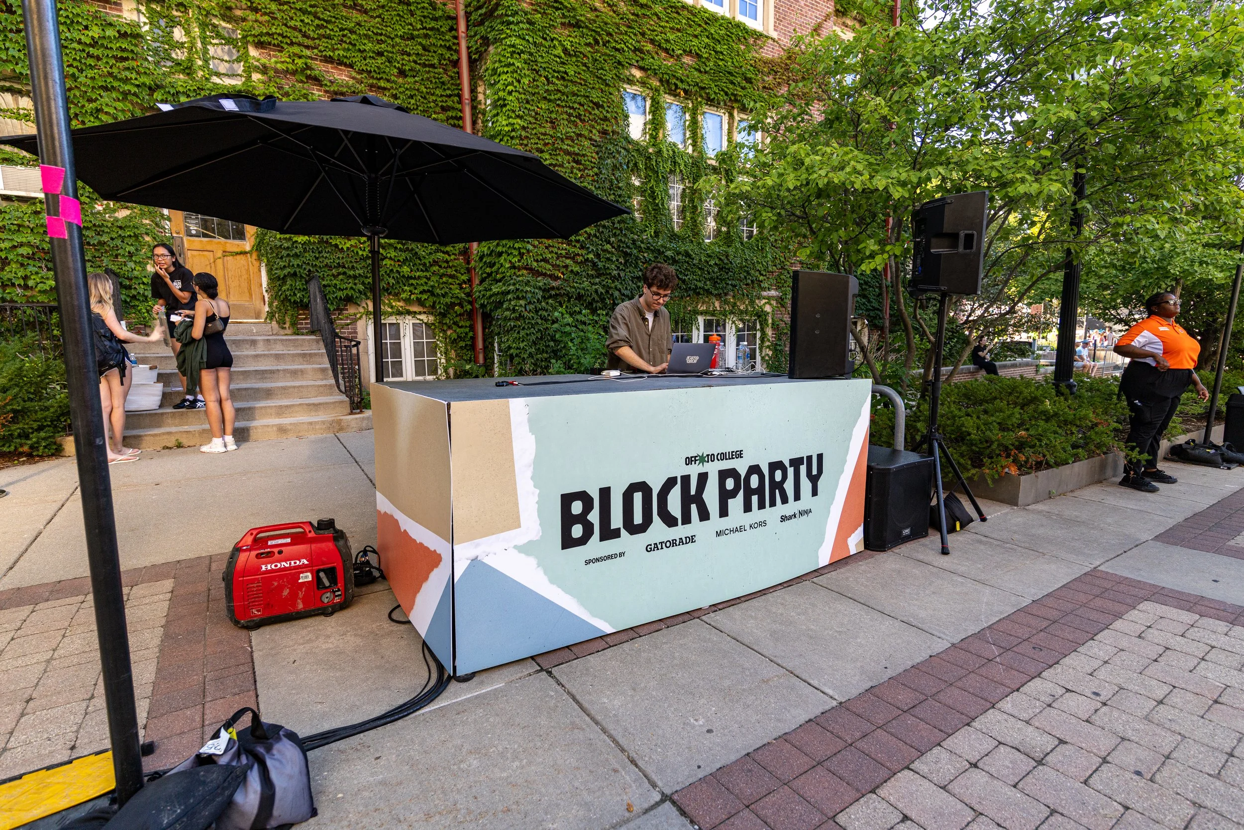 A DJ booth decorated for a block party with a sign that says 'Off to College Block Party' and a man working on a laptop, with people socializing nearby and a large black umbrella above.