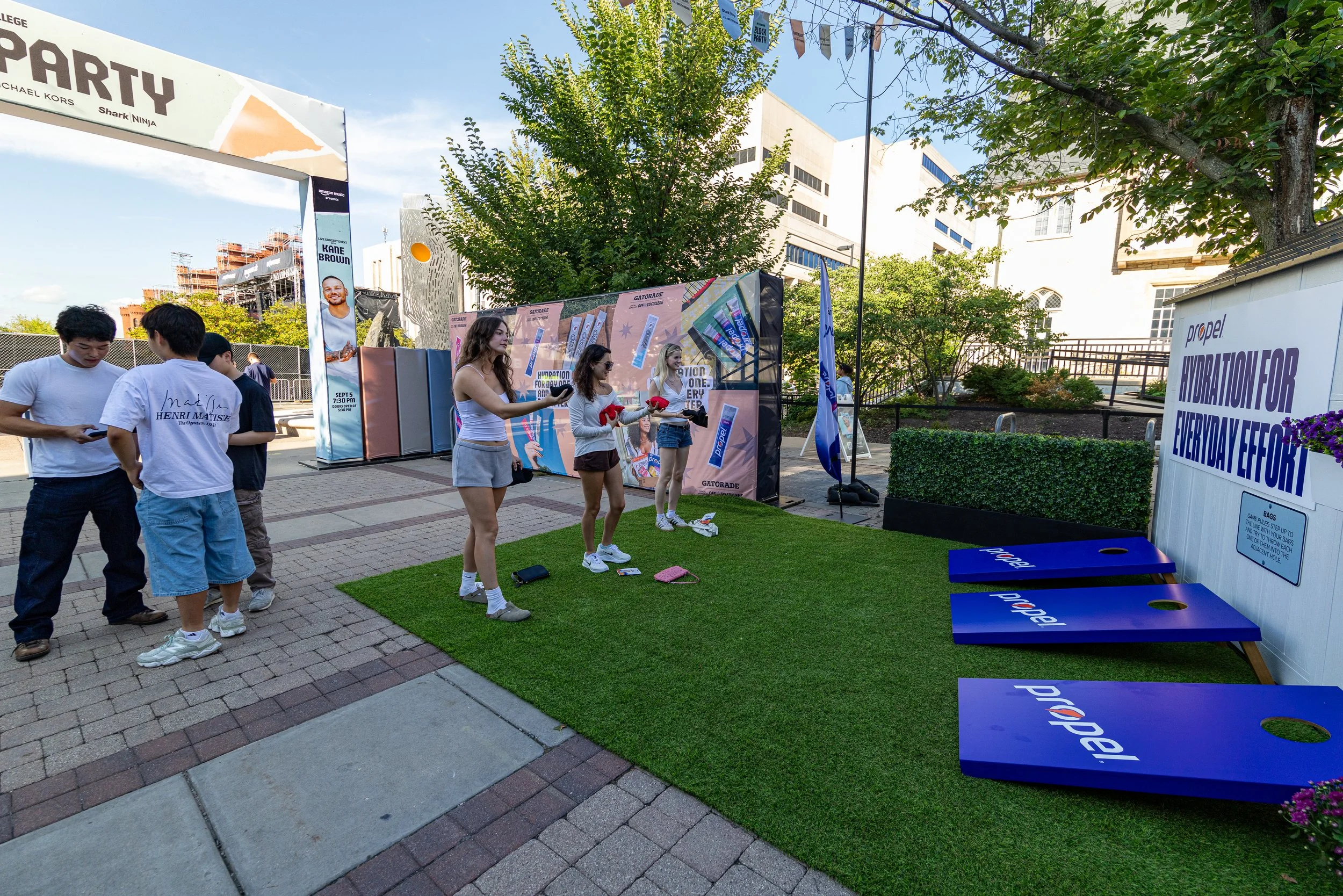 People participating in an outdoor event with games and activities, including a cornhole game with blue boards and a large sign for Propel water, with trees and buildings in the background.