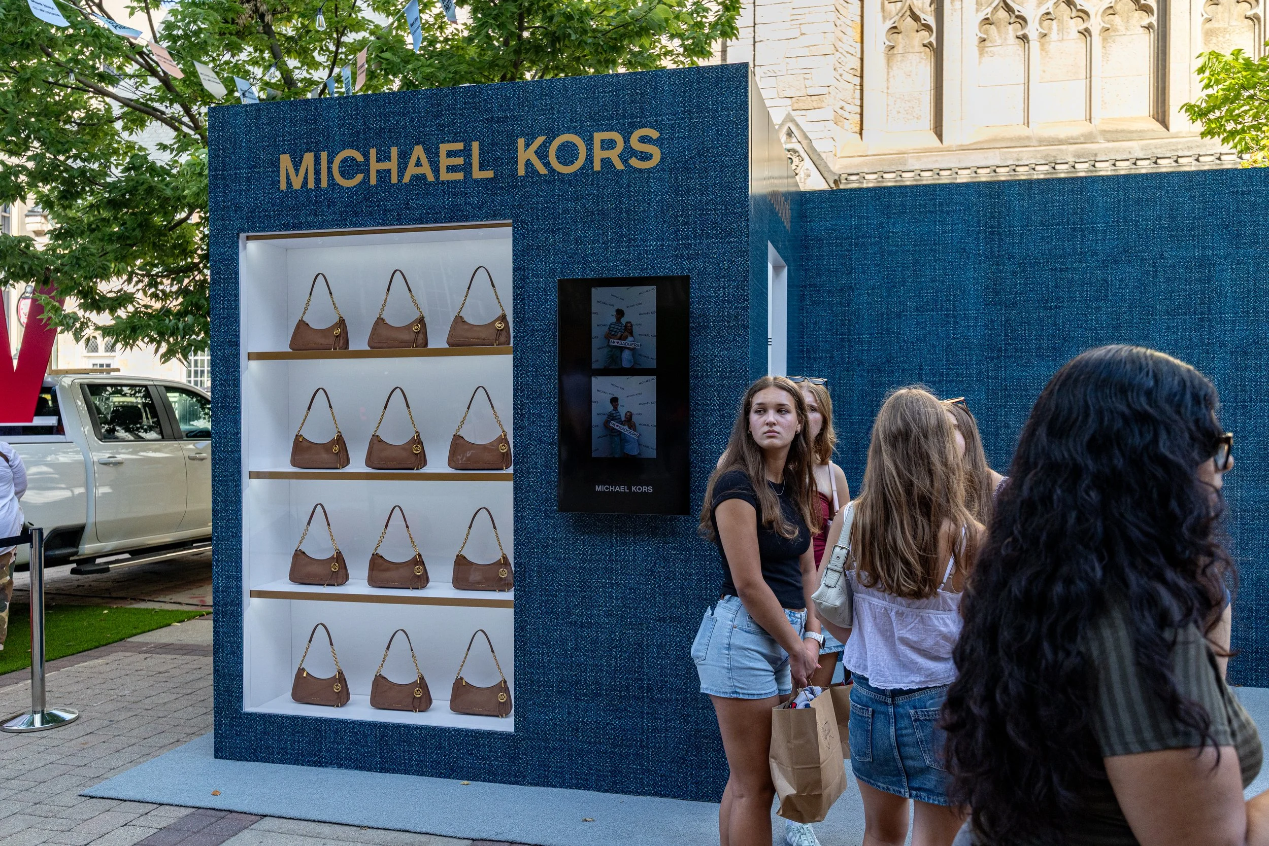 A Michael Kors display stand with designer handbags on shelves at an outdoor event, with multiple people standing nearby and trees in the background.