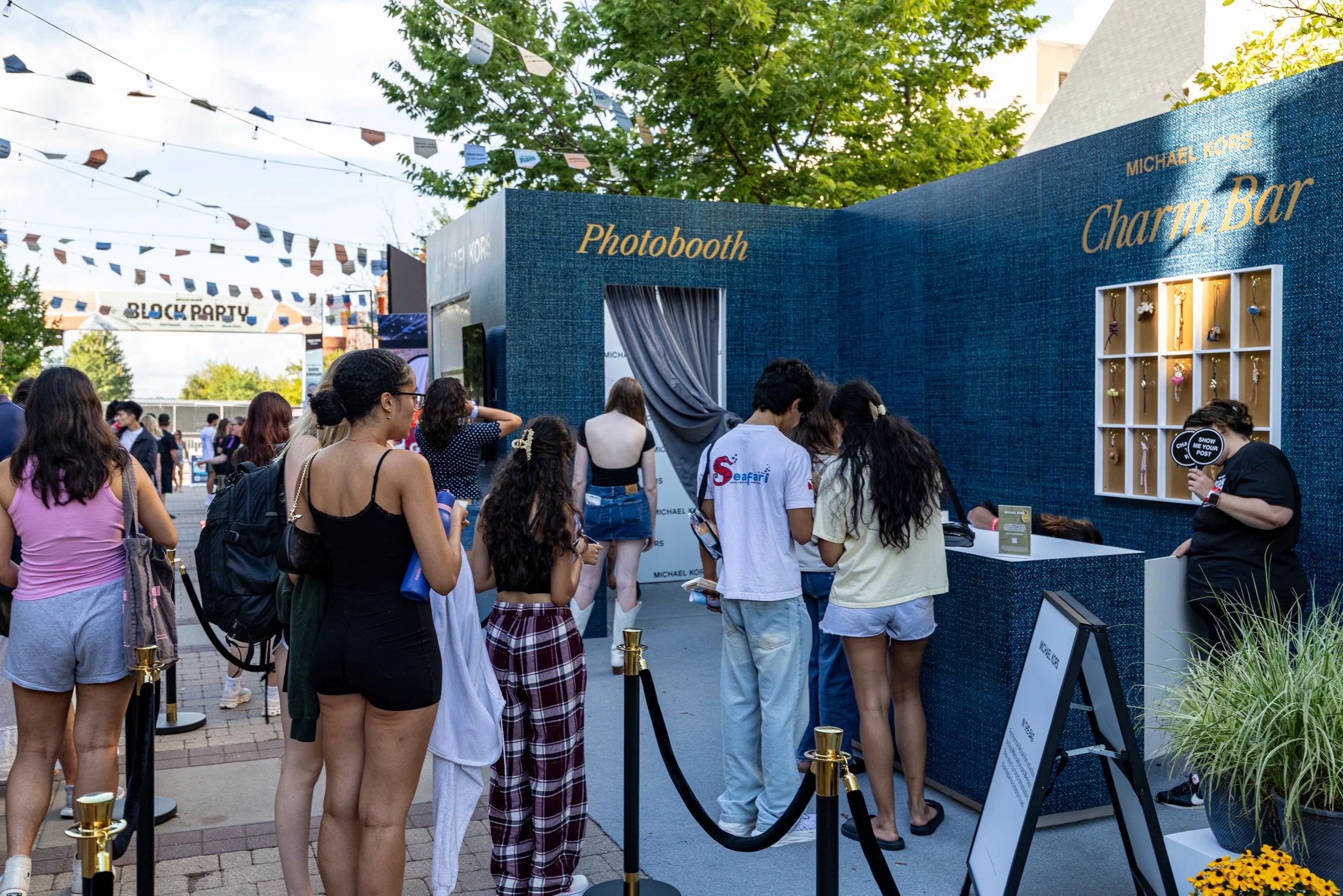 Line of people waiting at a Michael Kors Charm Bar photo booth at an outdoor event, with a blue textured wall backdrop and displays of jewelry, surrounded by trees and festoon lighting.