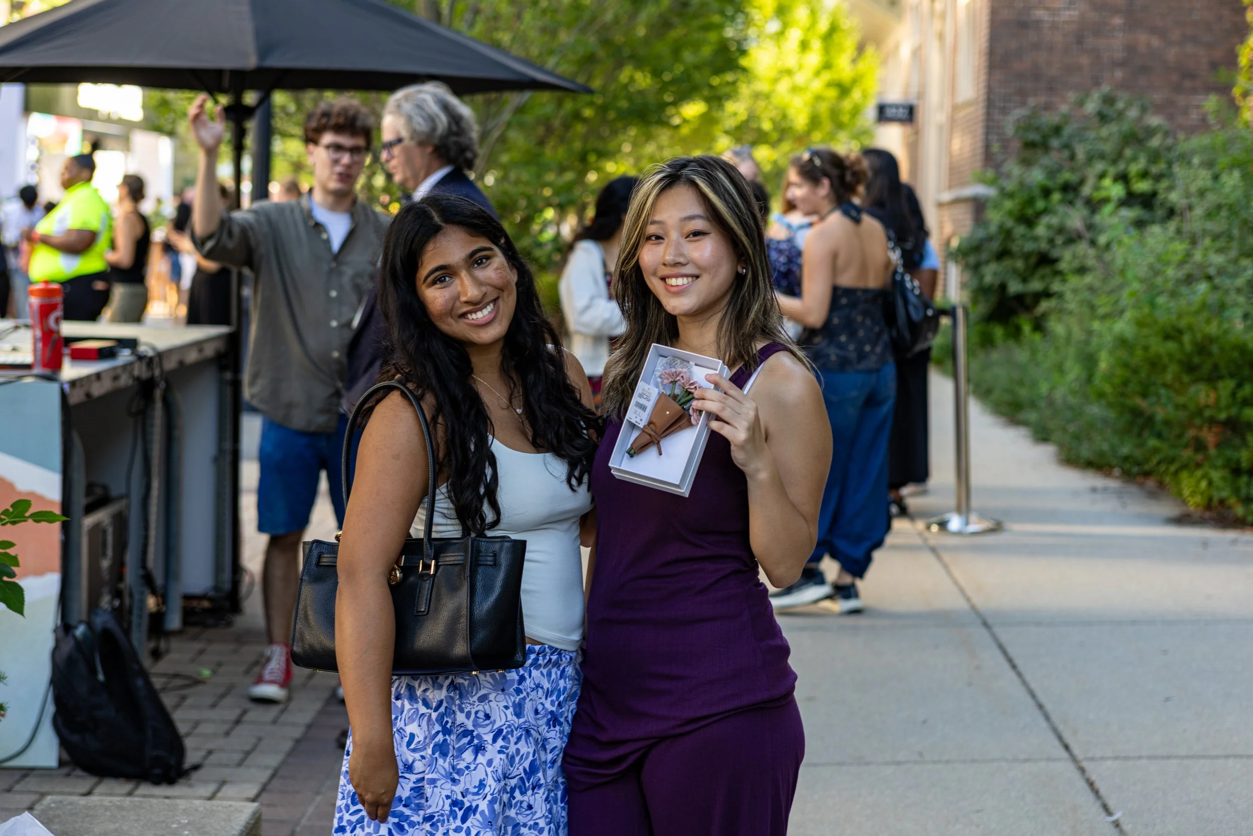 Two smiling women standing outside at an event, holding a box with a flower inside, with other people in the background.