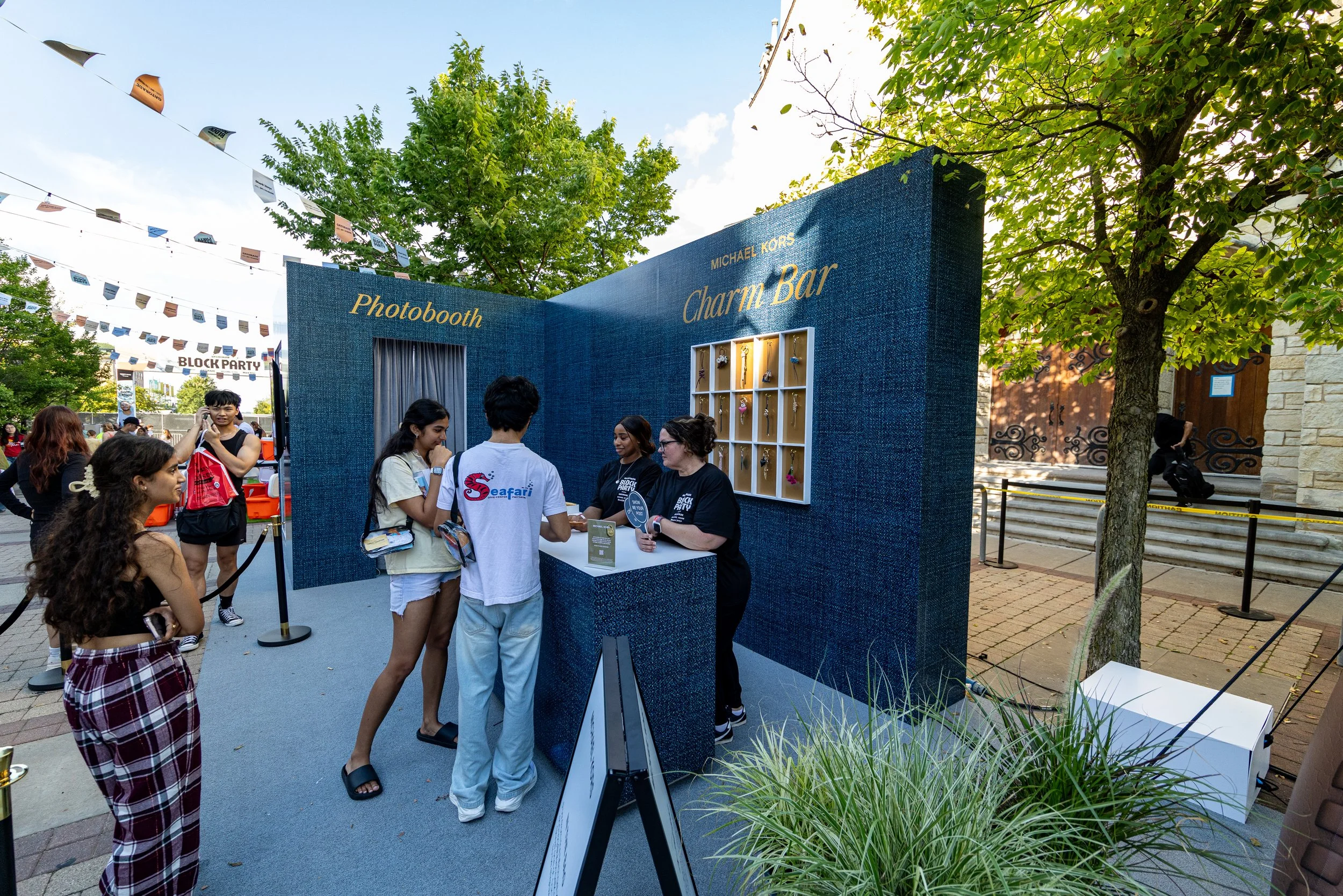 People gathered at a Michael Kors Charm Bar booth outdoors, with a blue textured wall, display shelves of charms, and a sign reading 'Photobooth.' Some are chatting with staff, and others wait in line.
