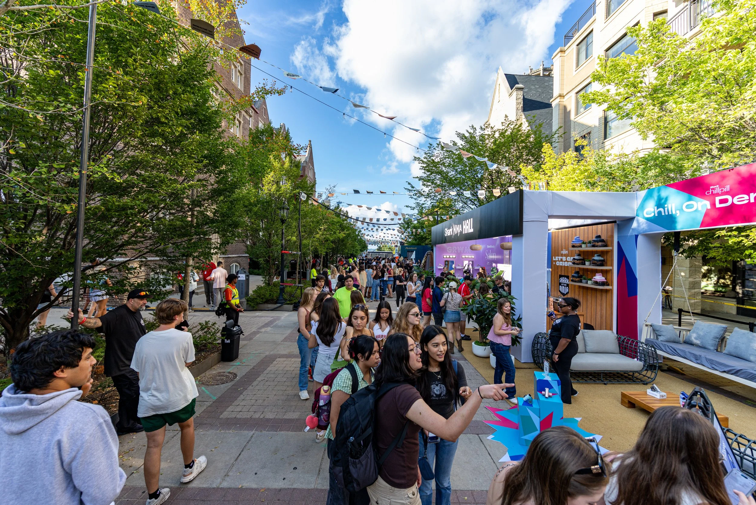 A lively outdoor event with a crowd of people walking along a tree-lined street under colorful banners. There are booths and tents, including one with a sign that says 'Chill. On D...'. The sky is partly cloudy and buildings are visible in the backgr