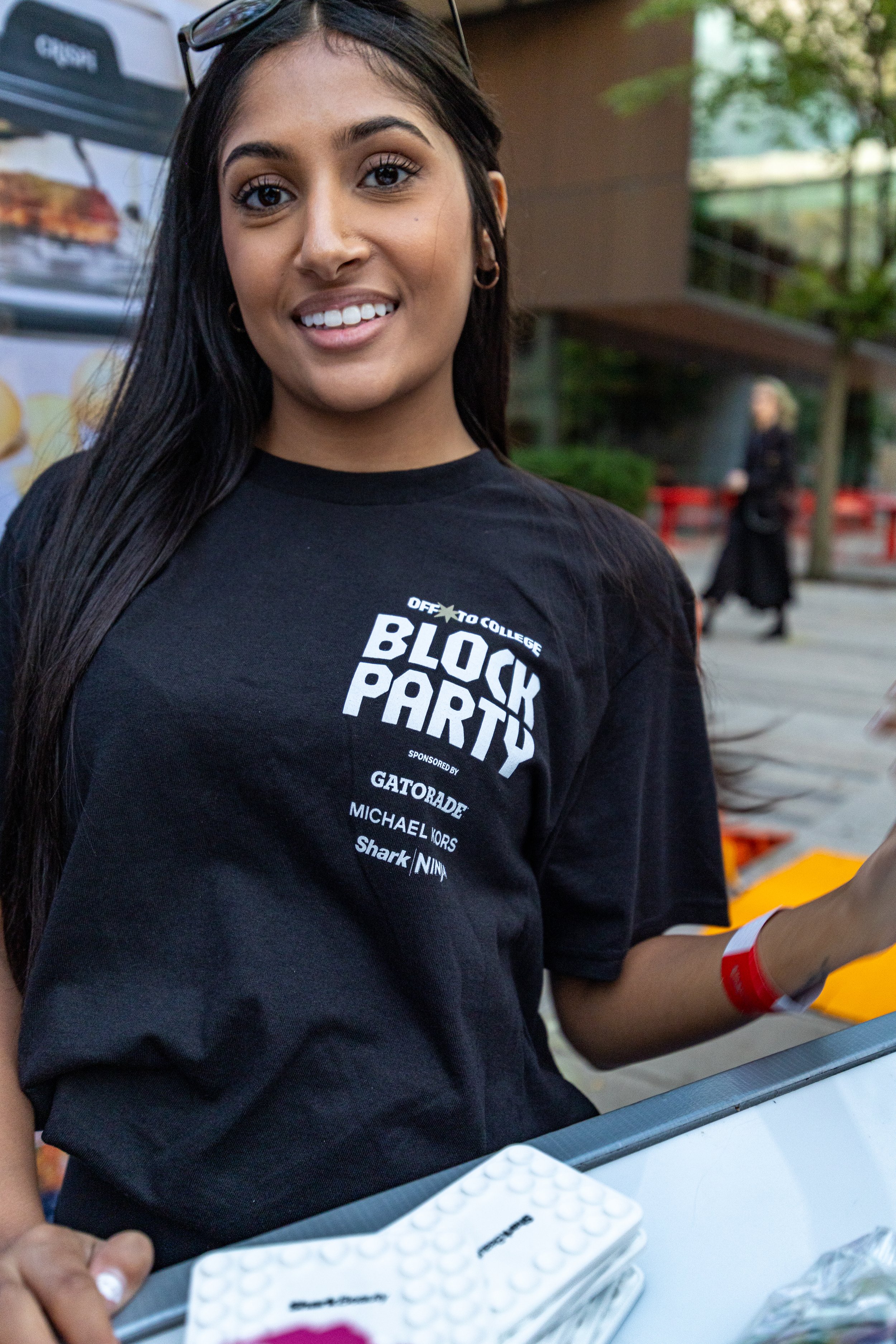 A young woman with long dark hair and wearing a black T-shirt that reads 'Block Party' is smiling at the camera. She has sunglasses on her head and small hoop earrings. She is standing outdoors next to a table, possibly at an event or gathering.