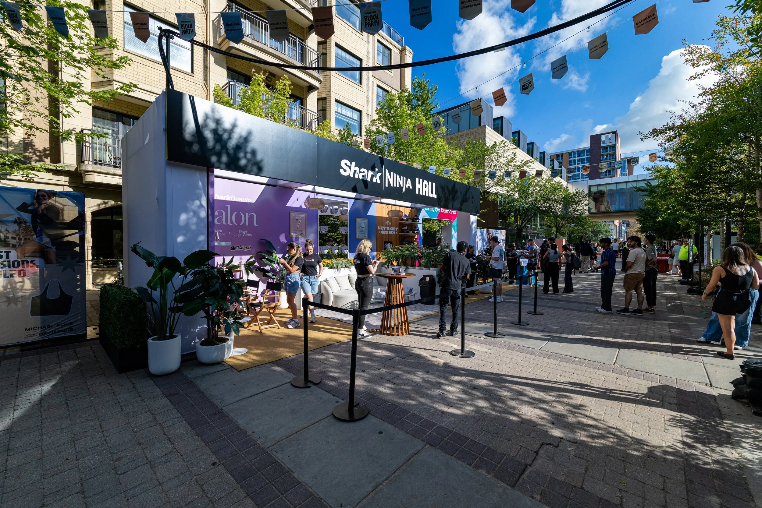An outdoor event setup with a stage or booth titled 'Shark Ninja Hall', surrounded by people, banners hanging above, and trees in an urban area under a partly cloudy blue sky.