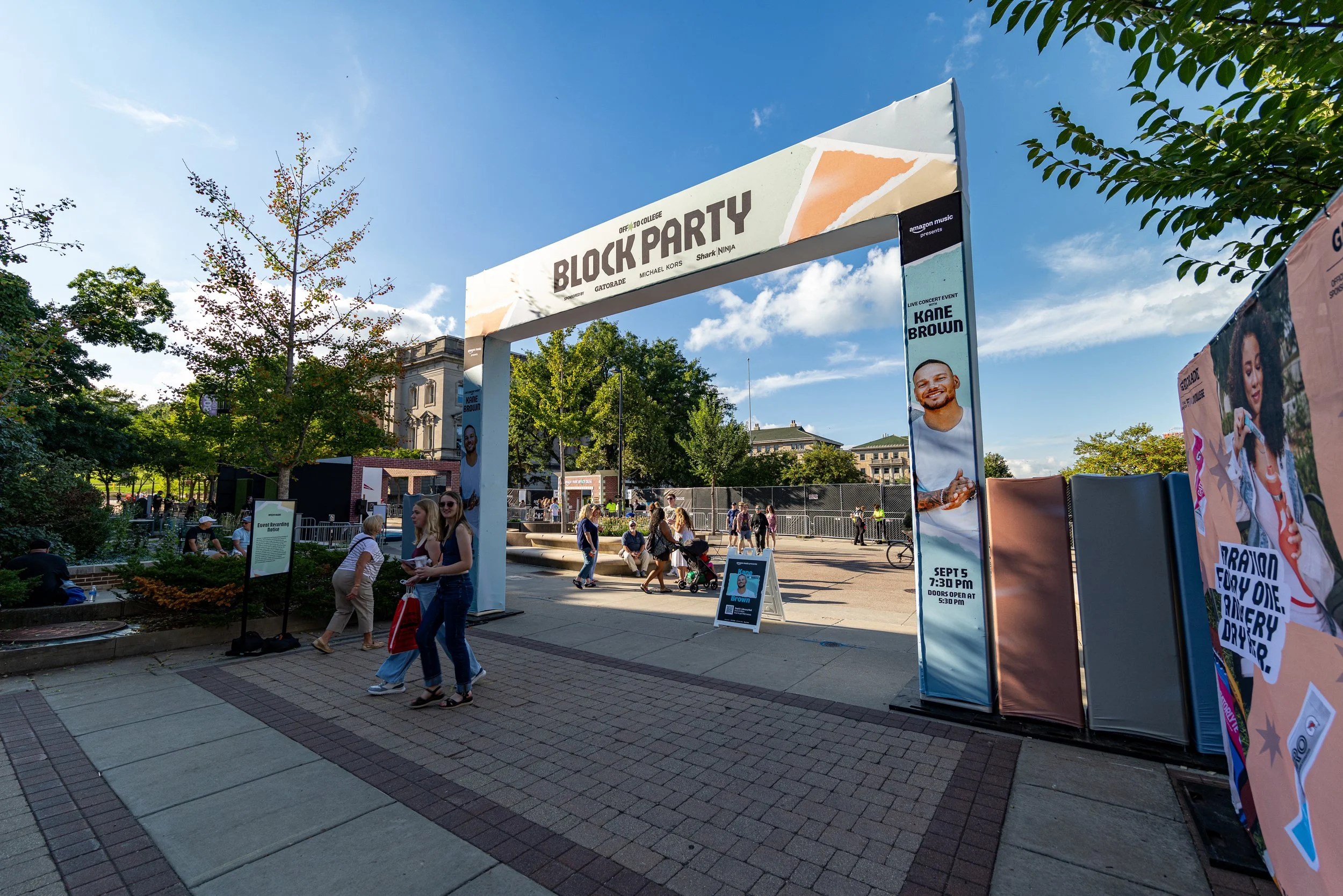 Outdoor event area with a large entrance archway promoting a Kane Brown concert on September 5th at 7:30 PM, with attendees walking and sitting in a park-like setting under a blue sky.