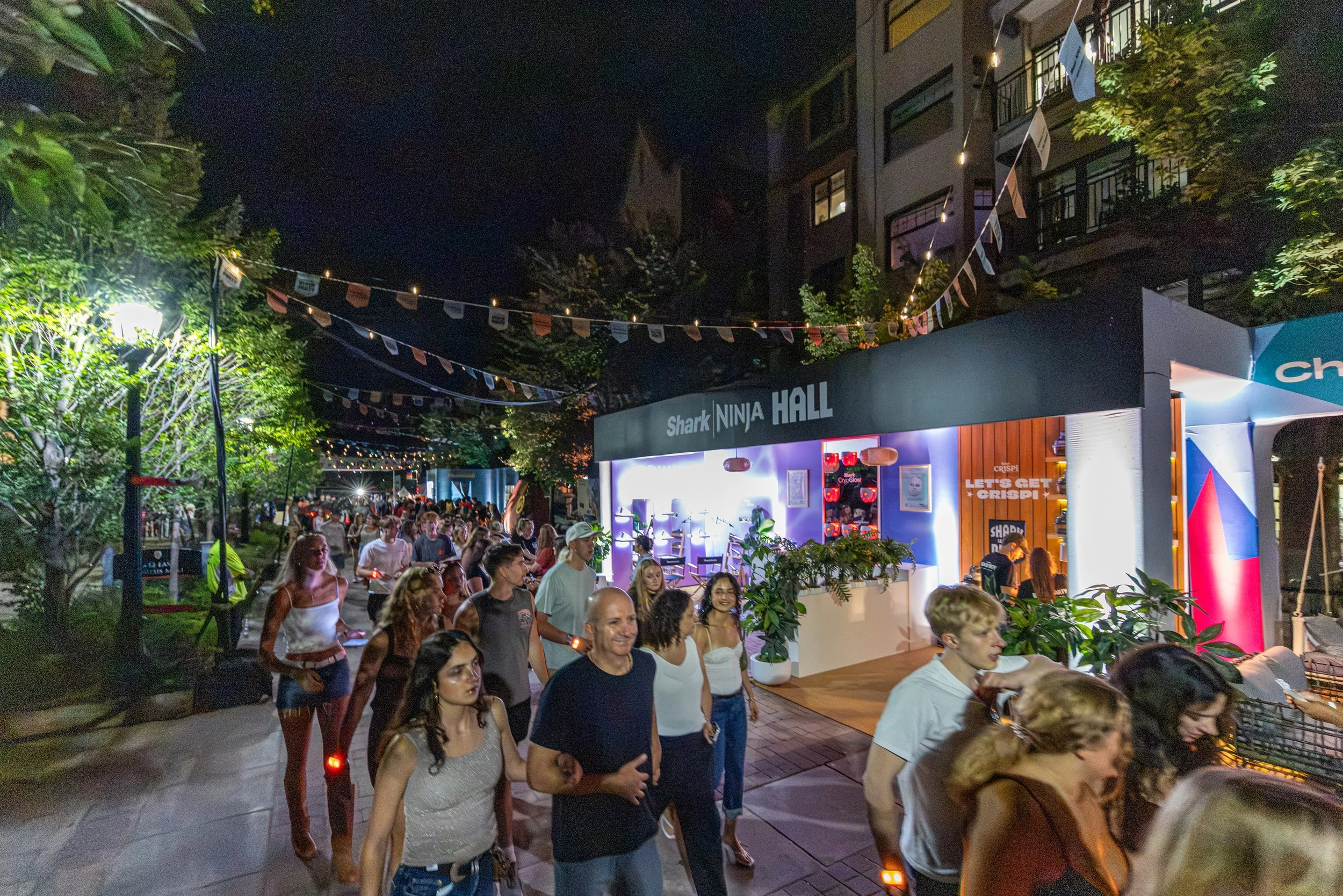 Crowd of people at night in an outdoor event with trees, string lights, and a building with balconies in the background. There are party decorations and a booth labeled "SHARK NINJA HALL."