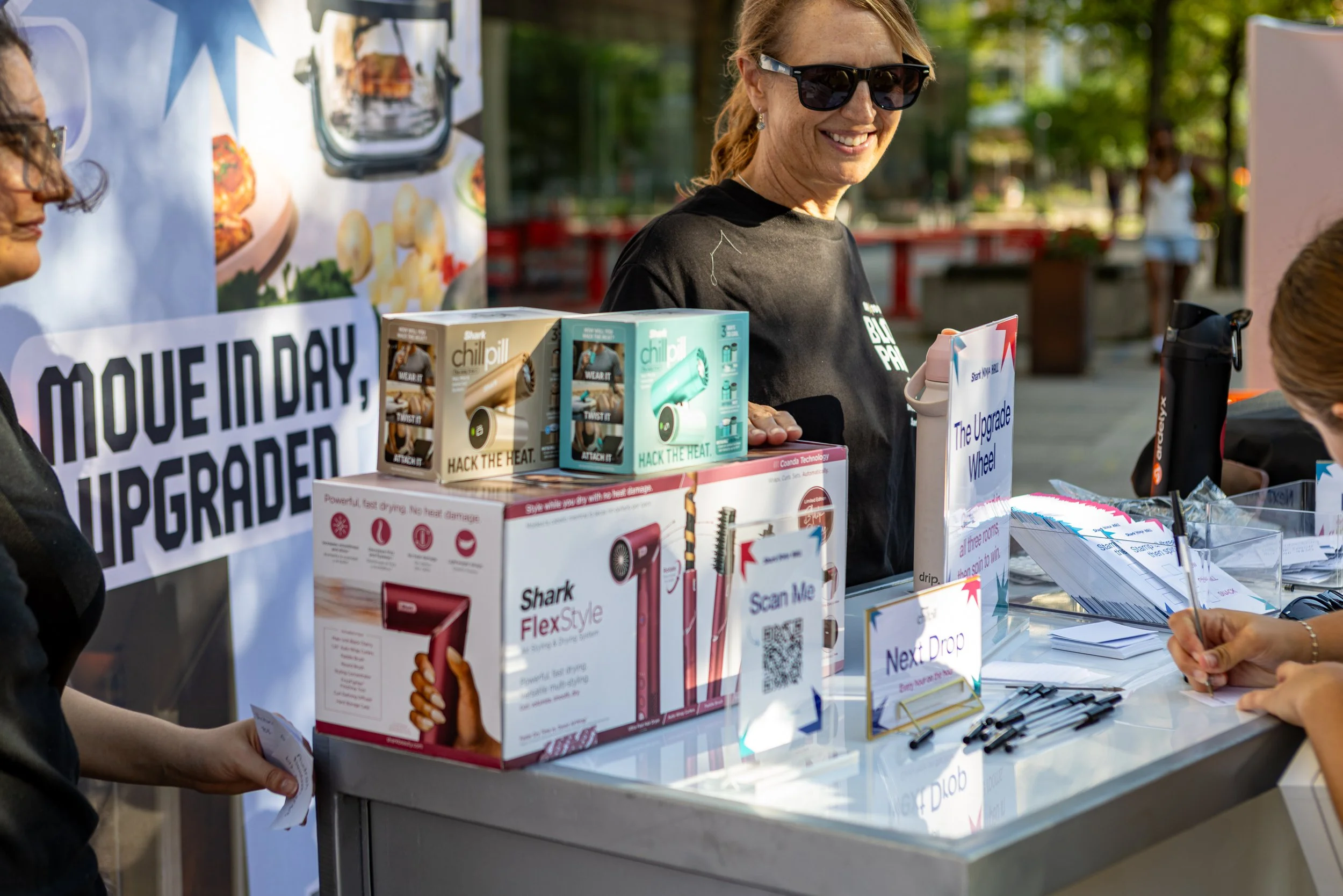 A woman in sunglasses smiling at a booth with various products and signs at an outdoor event.