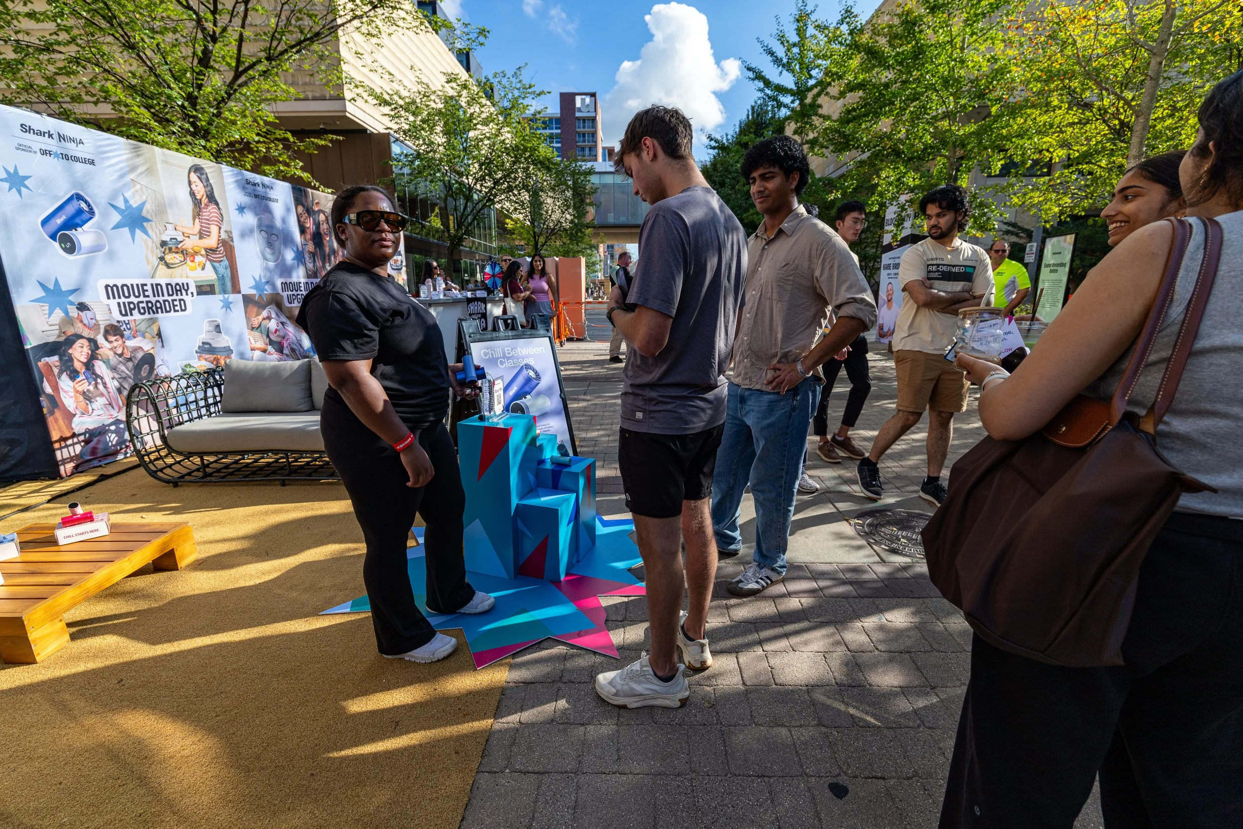 People gathered at an outdoor booth with banners promoting tech products, surrounded by trees and city buildings, on a sunny day.