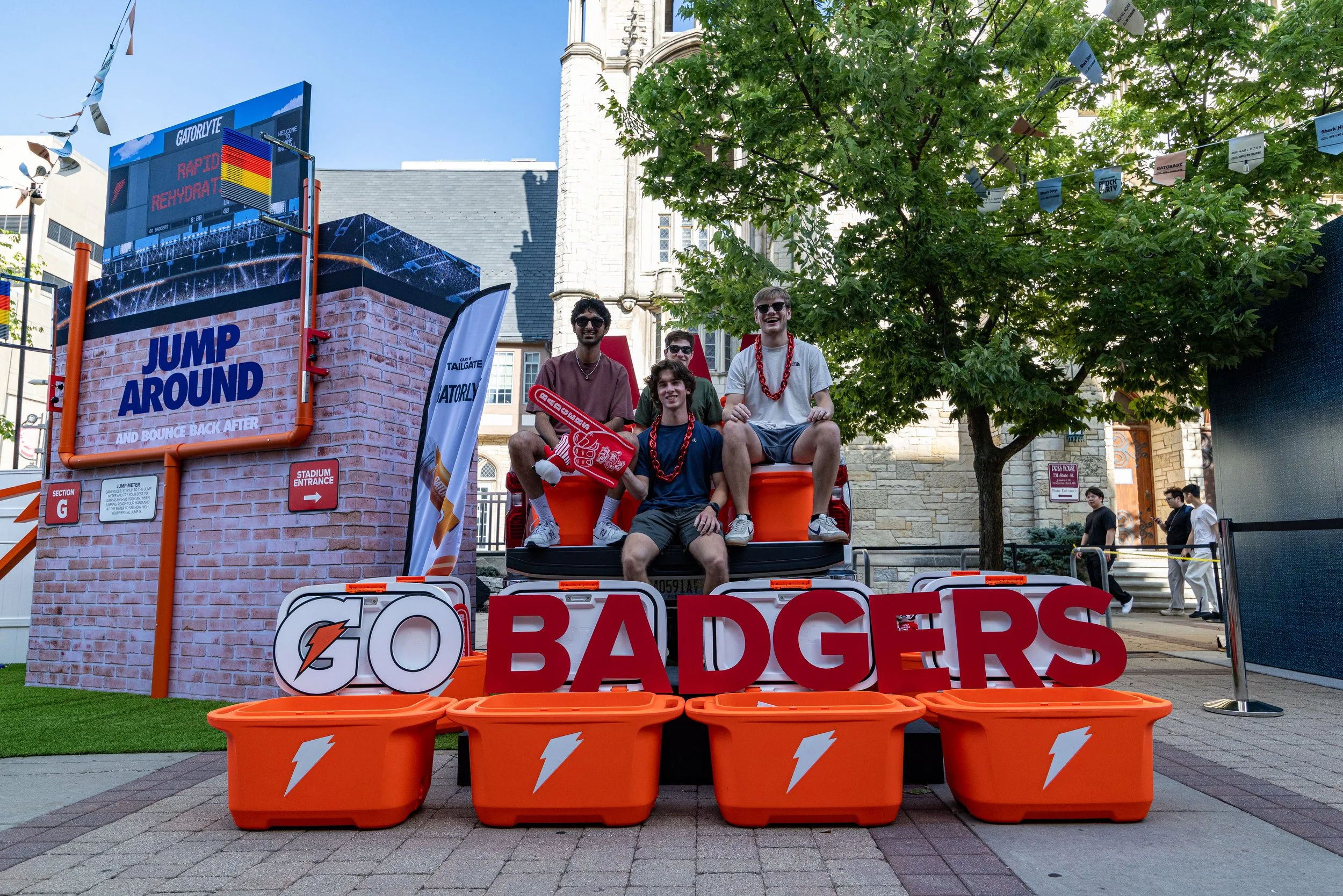 A group of five young men sitting on a small orange vehicle with orange buckets in front, decorated with sports-themed accessories, at an outdoor event near a large tree and a historic stone building, with signs and banners related to the event.