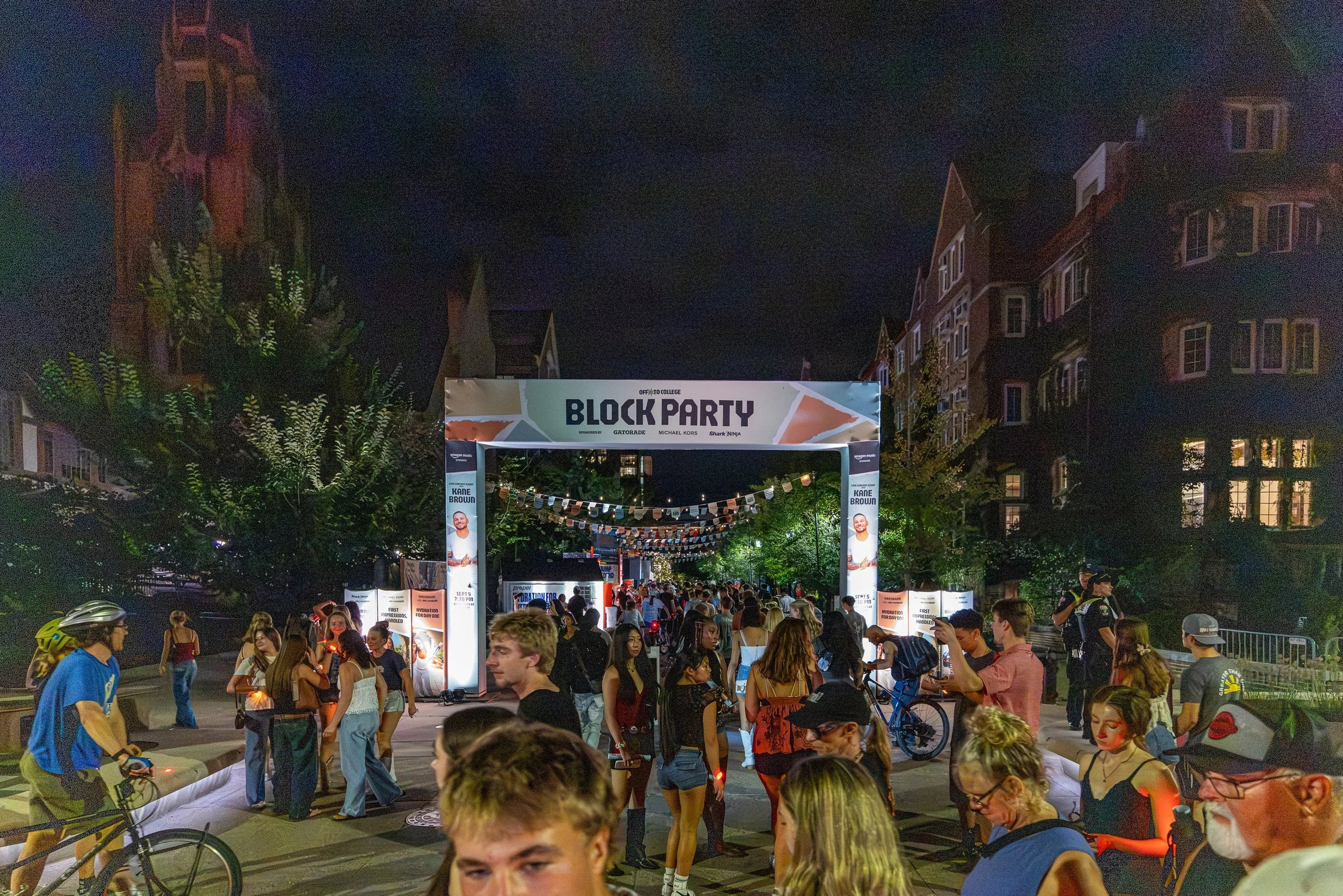 People attending a night outdoor event called 'Block Party' with a large archway entrance and string lights, surrounded by city buildings and trees.