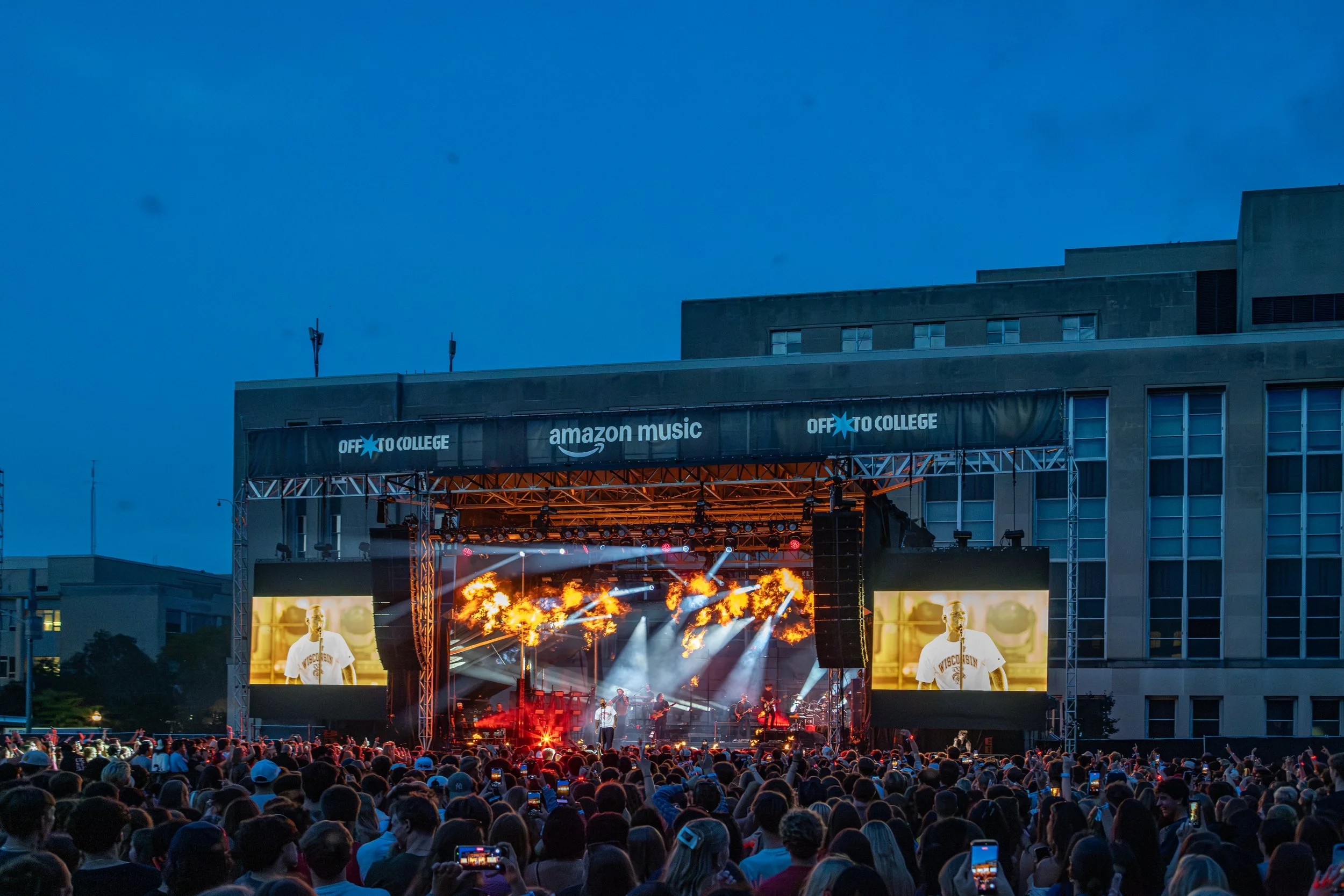 An outdoor concert at dusk with a crowd in front of a stage, bright lights, flames, and a large screen displaying a performer wearing a University of Wisconsin shirt.