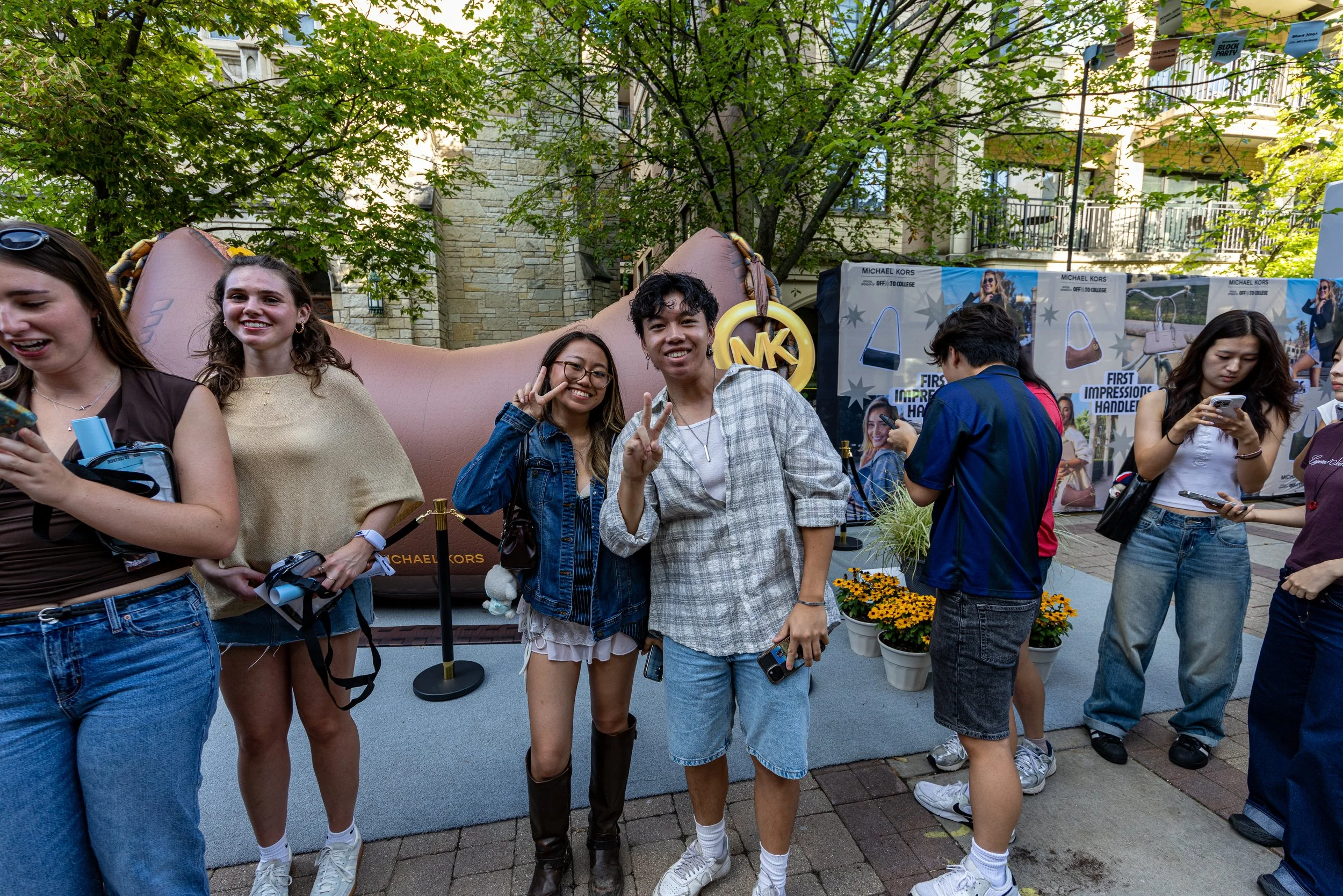 Group of young people outdoors at an event, with some smiling and making peace signs, others looking at their phones, and a backdrop advertising a Michael Kors display.