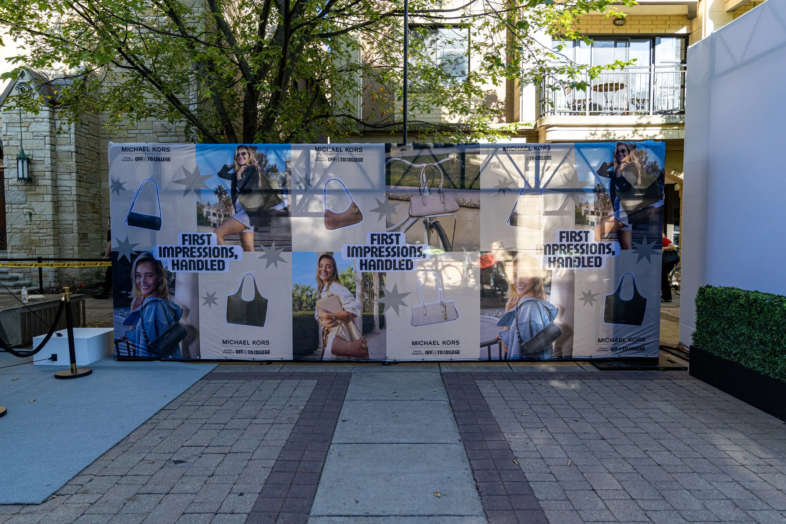 Outdoor display with Michael Kors advertisements featuring women posing with handbags, trees, and a building in the background.