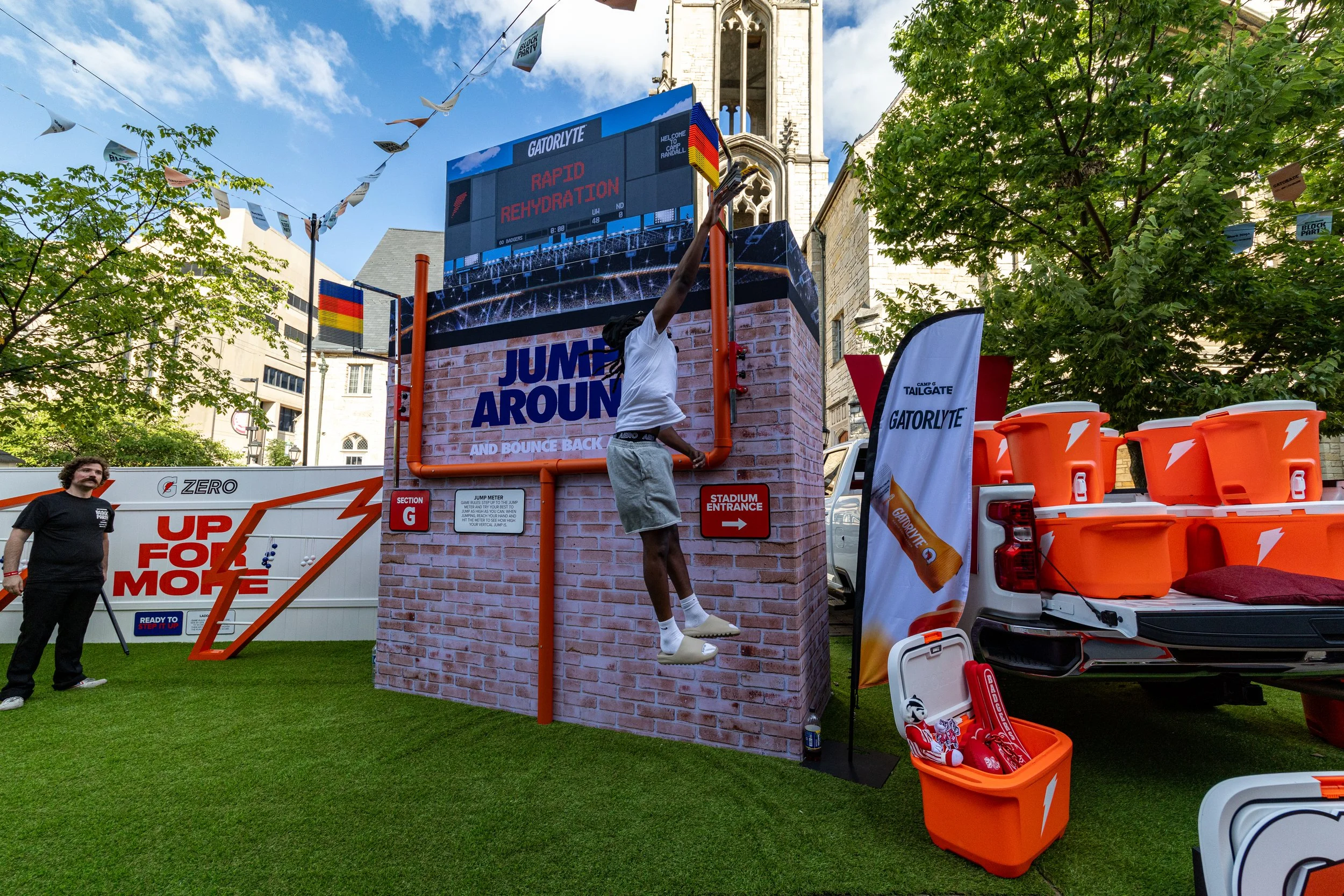 Person jumping to grab a basketball at a sports event with a large digital scoreboard displaying 'Rapid Rehydration.' There are trees, flags, and event banners, including Gatorade branding, in an outdoor setting with a historic building in the backgr