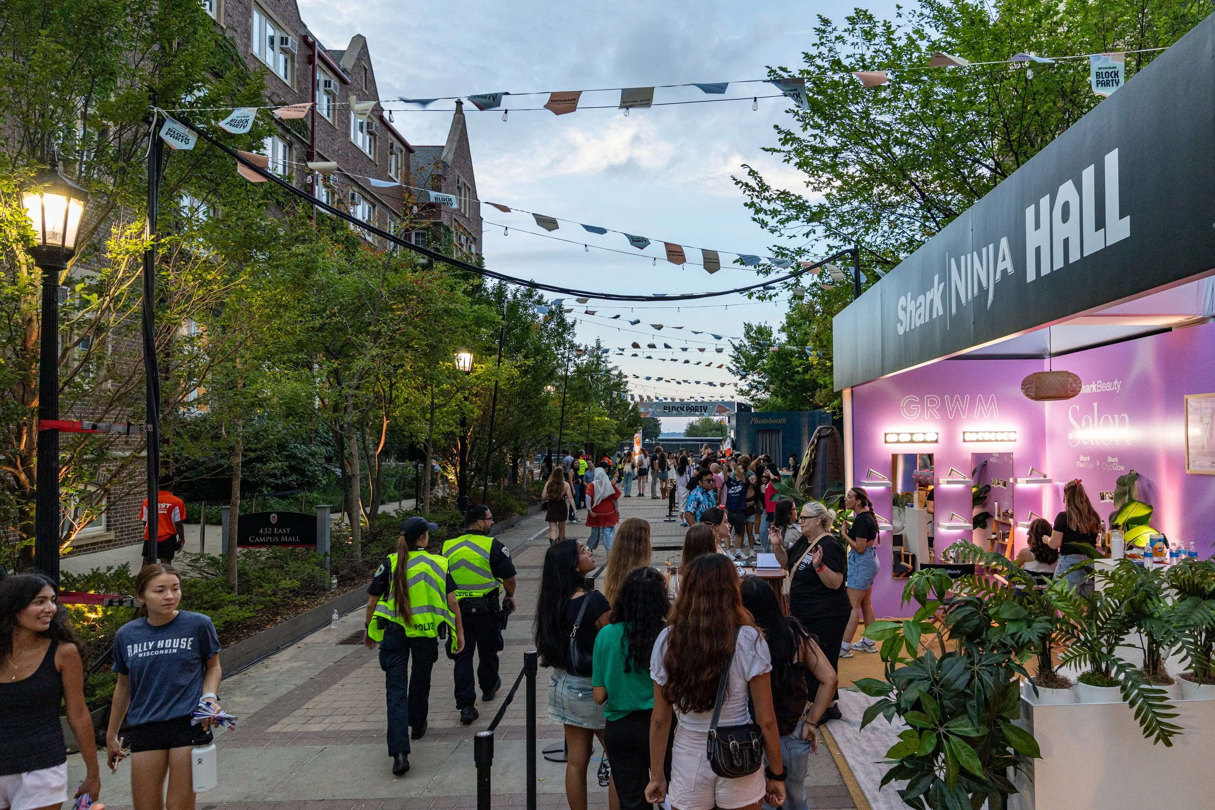 People attending an outdoor event or festival on a city street, with vendor booths and string lights overhead, and some people wearing yellow police vests.
