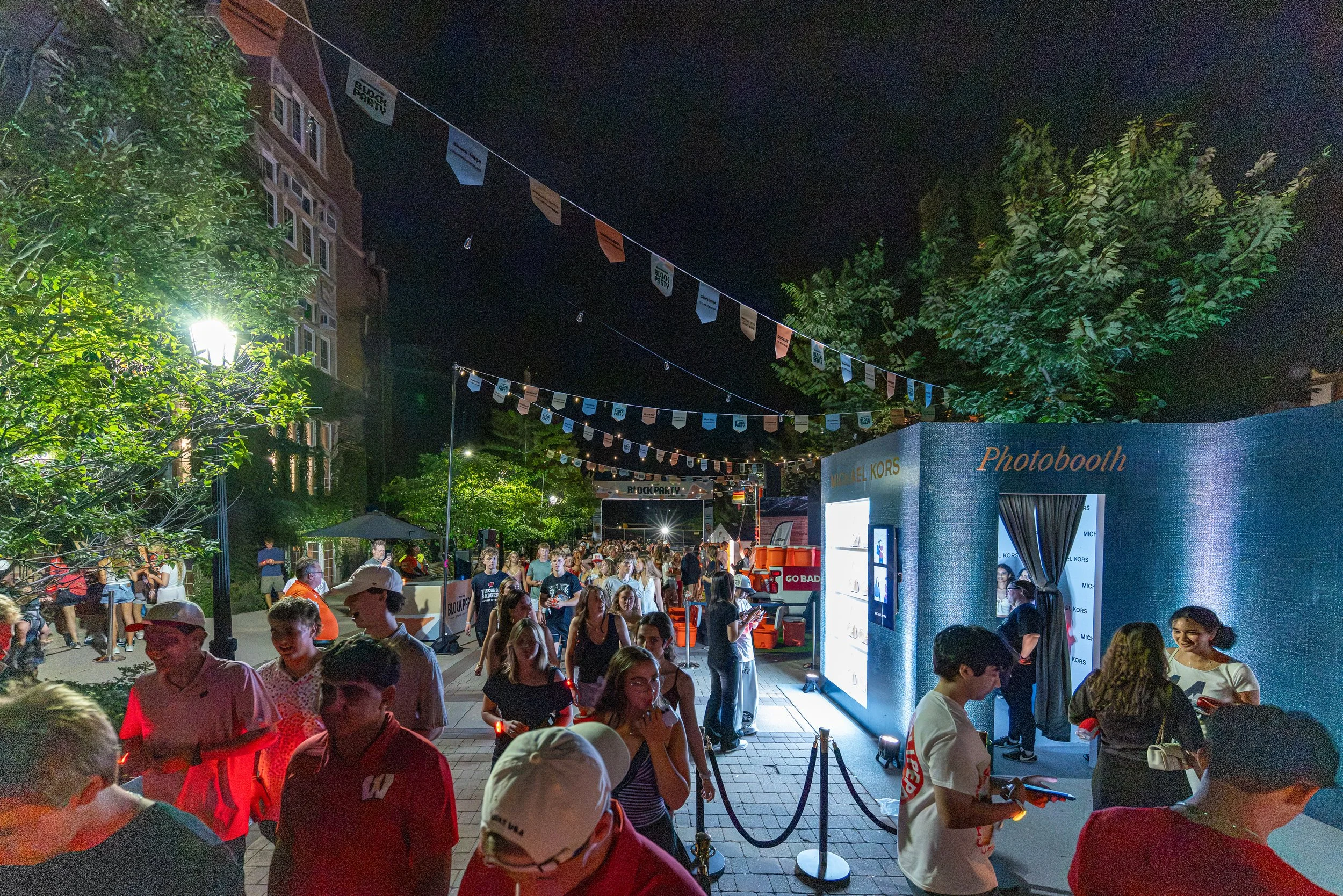 Night scene of an outdoor event with a crowd of people, string lights, trees, and a photo booth booth with signs, as people gather and socialize.