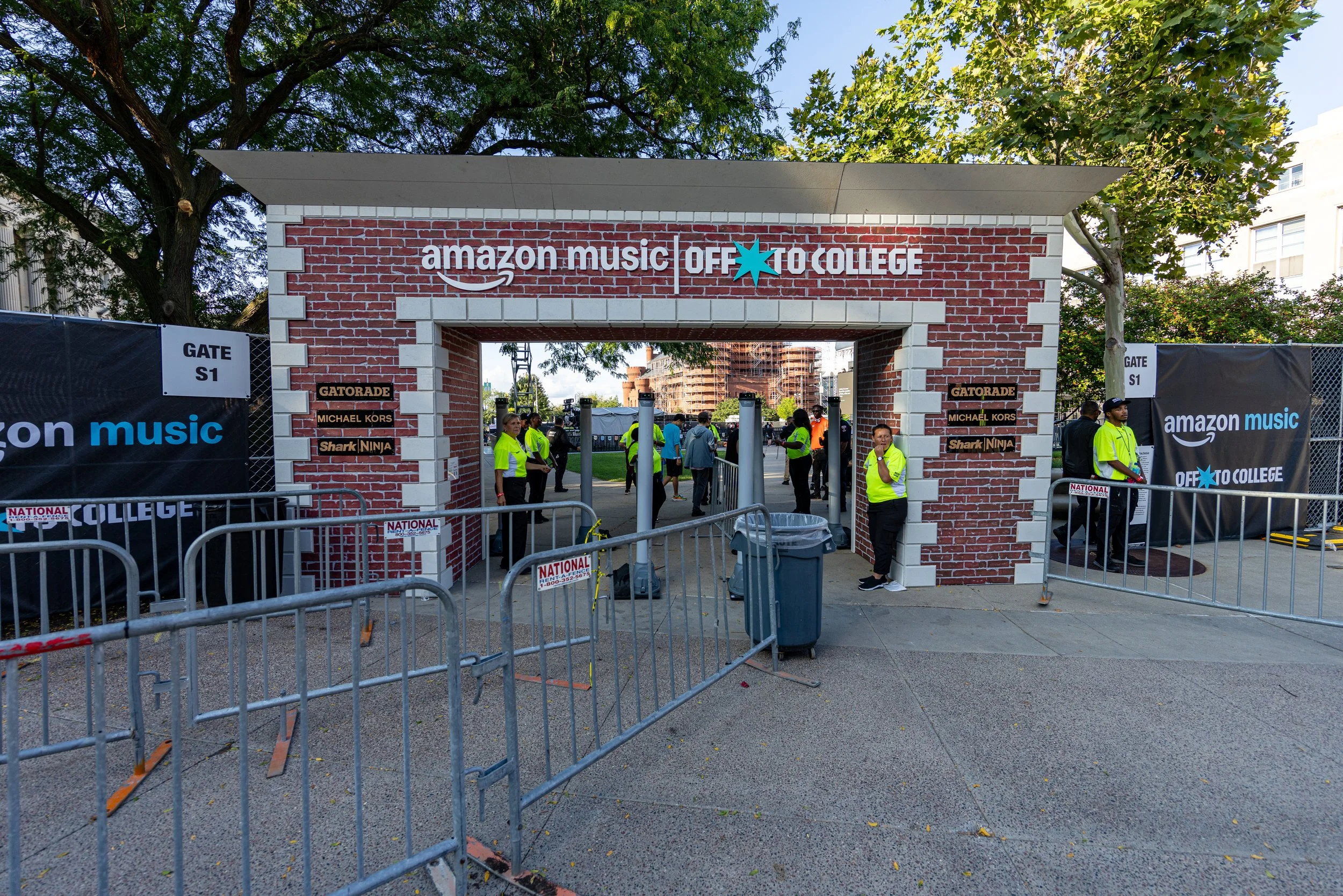 Entrance to an event with a brick archway that reads 'amazon music OFF TO COLLEGE'. Security personnel and event staff are present, with barriers set up and banners displaying partnerships with 'Gatorade', 'Michael Kors', and 'Shark Ninja'. Trees and