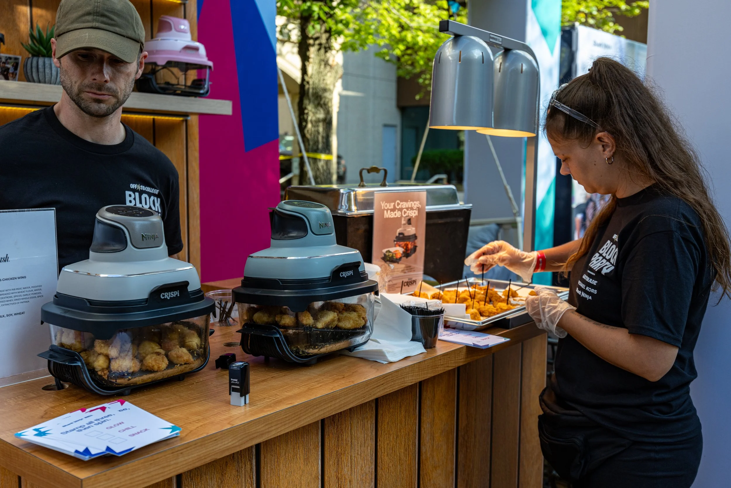 A food stand at an outdoor event with two staff members preparing food. The woman is wearing gloves and working on a tray of fried food, while the man looks down at the counter. There are food warmers with fried items on the wooden counter and a colo