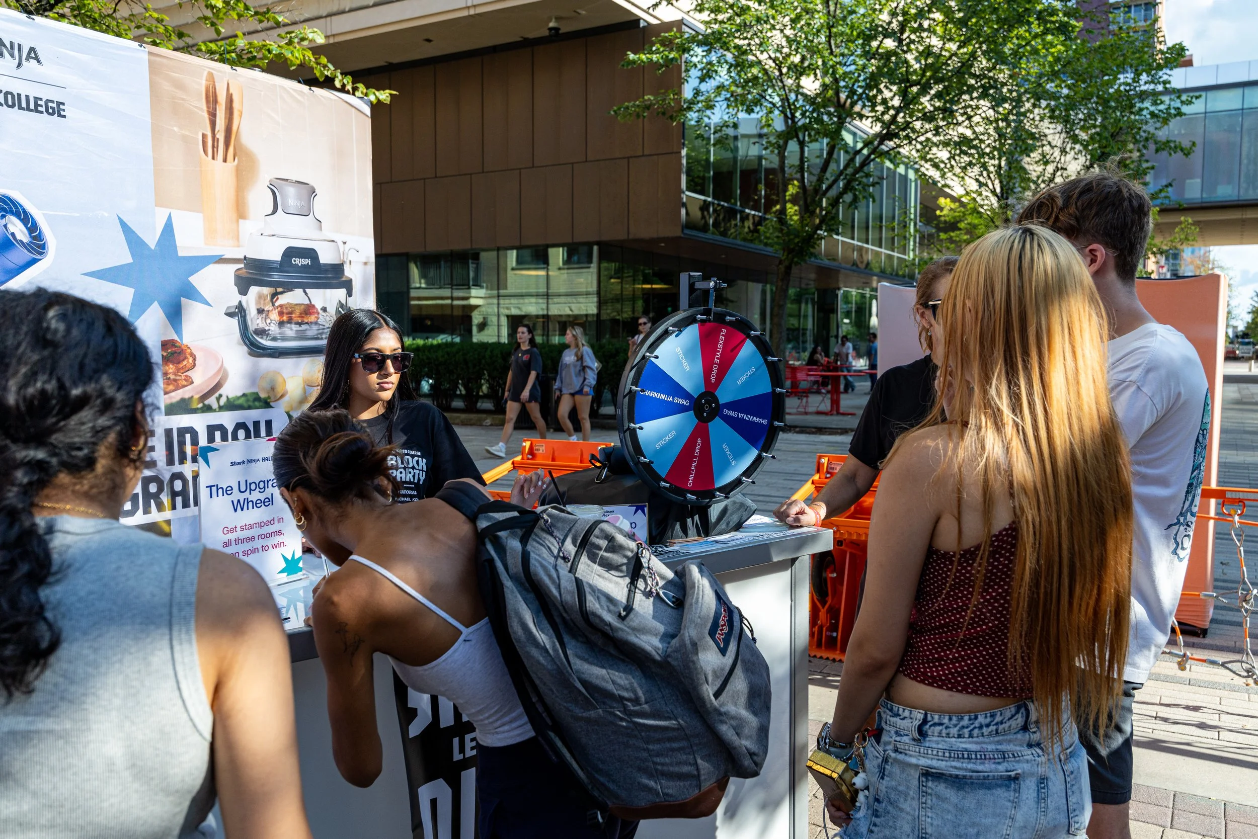 A group of people gathered at an outdoor booth with a spinning prize wheel, likely at a street fair or event, with trees and modern buildings in the background.