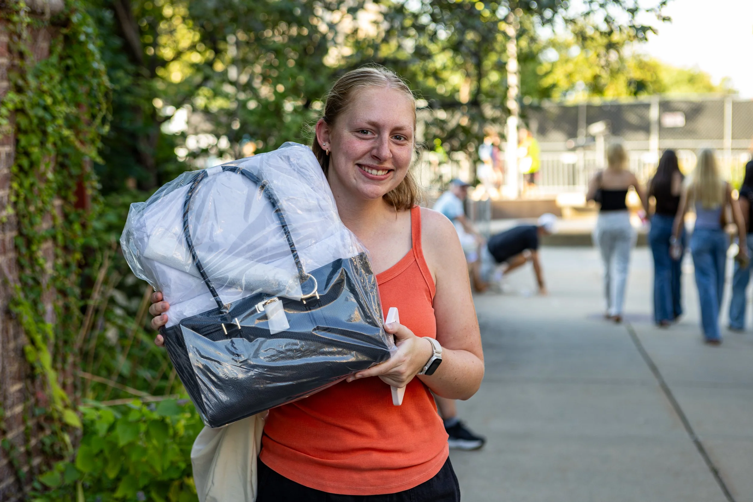 A young woman smiling and holding a large plastic bag with unknown contents in an outdoor park area with other people in the background.