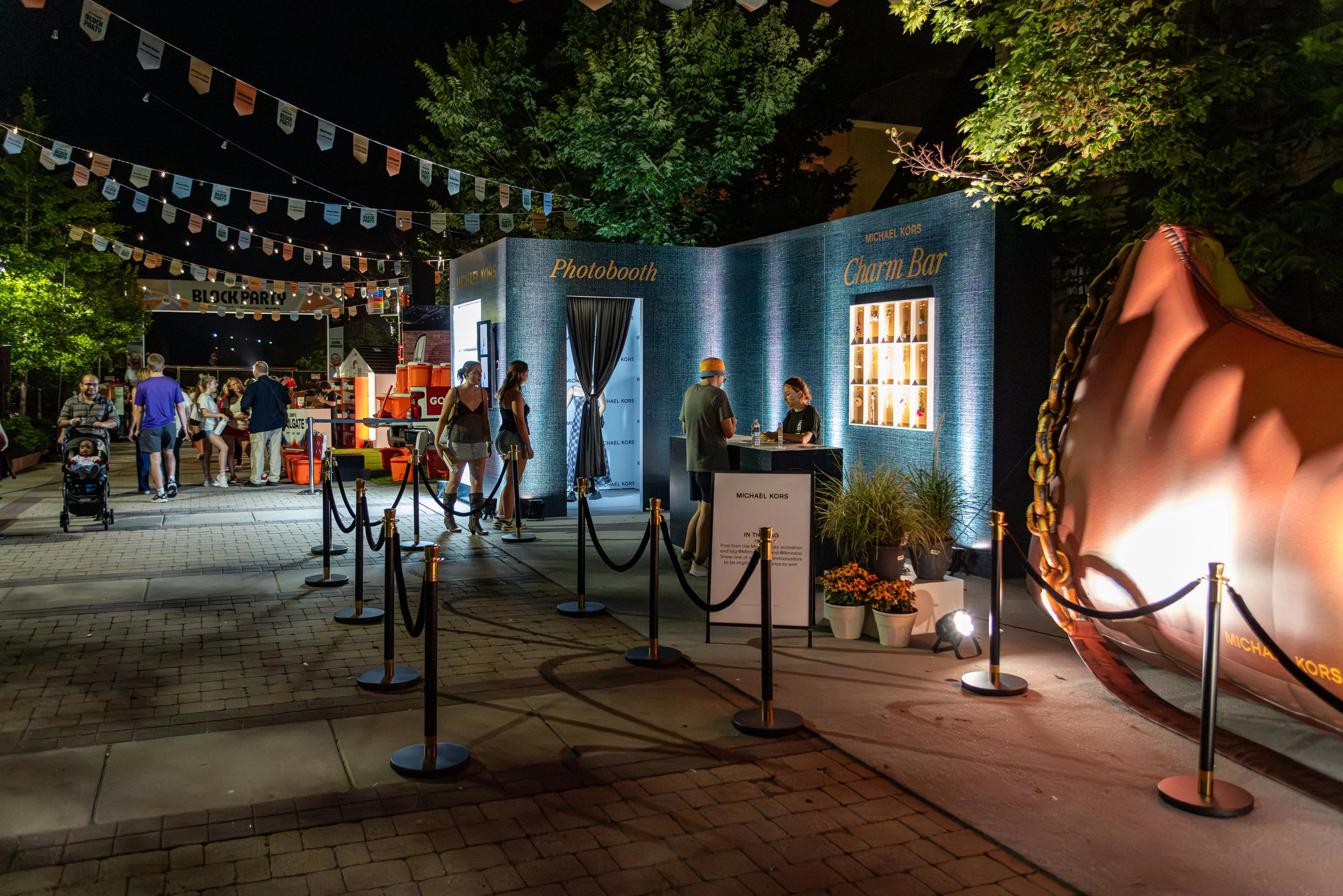 Nighttime scene of an outdoor event with people, food stands, and a photo booth area. String lights and banners are hanging overhead, and there are trees in the background. A large Michael Kors charm decoration is visible on the right.