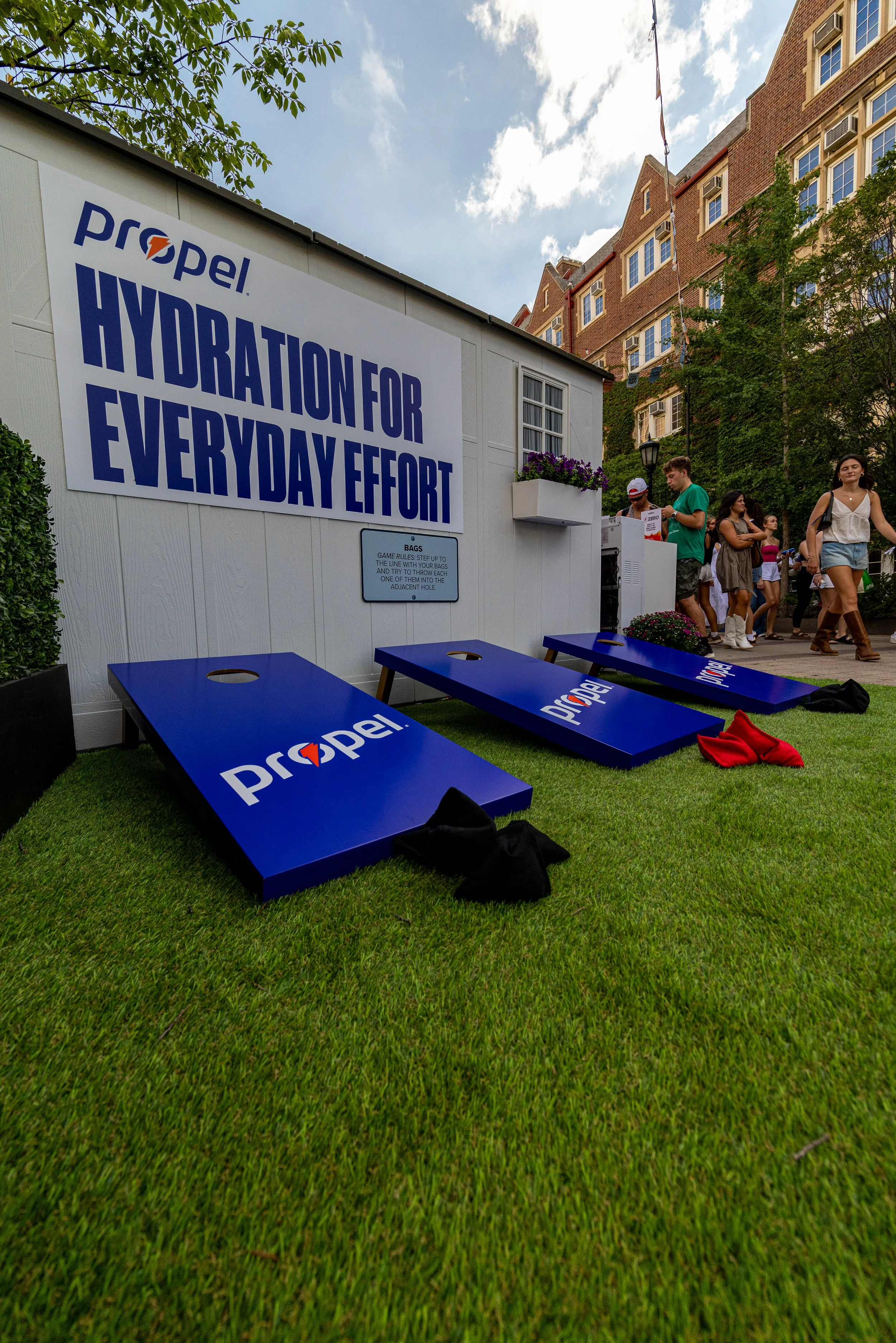Outdoor scene at a promotional event with giant blue cornhole game boards displaying 'Propel'. A white building with a large sign saying 'Hydration for Every Day Effort' is visible, as well as a group of people standing and walking on the sidewalk. T