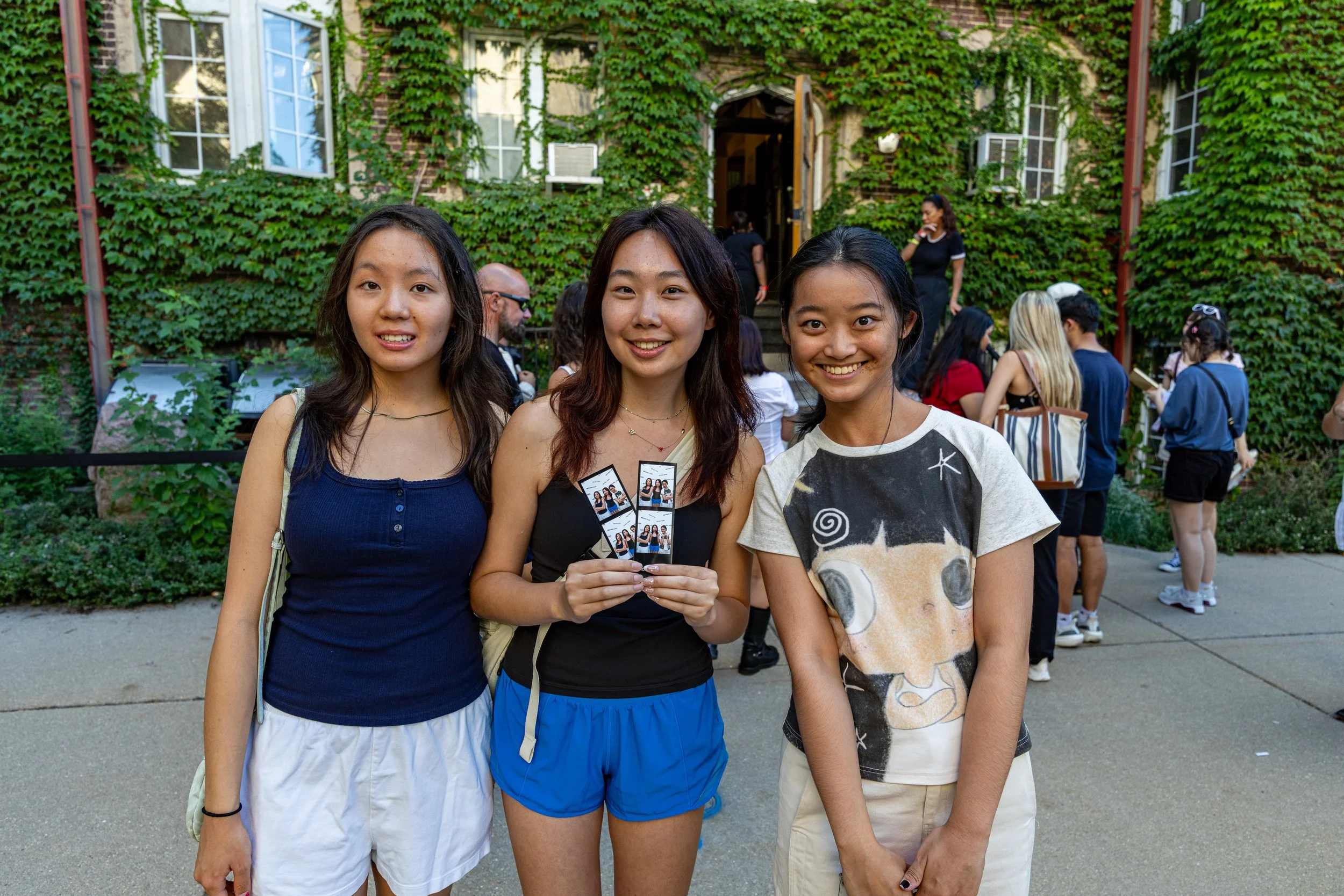 Three young women smiling and holding a group photo, standing outdoors in front of a green ivy-covered building with several other people in the background.