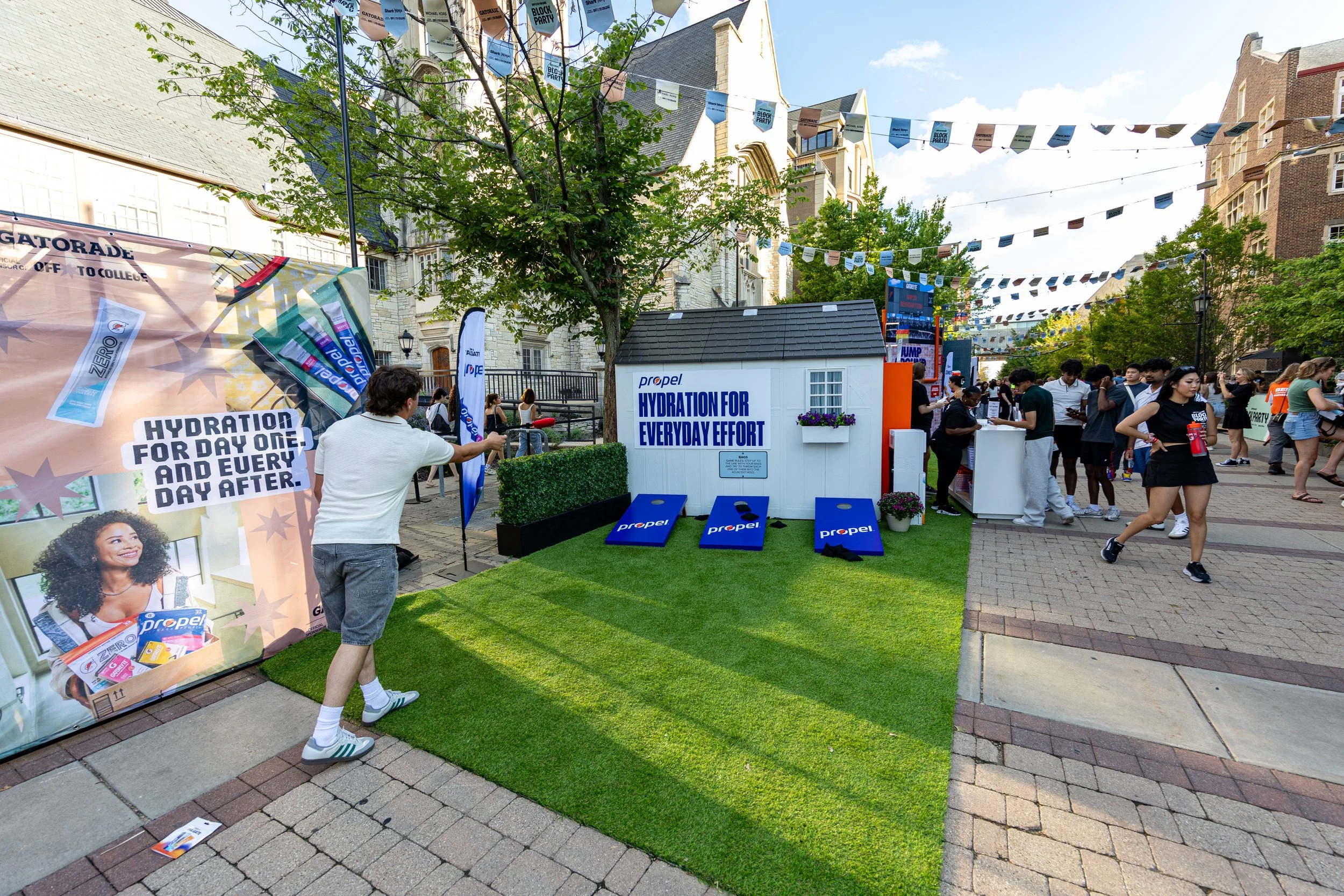 An outdoor event with people gathered around booths and displays promoting hydration products, with banners, informational signs, and a small house-shaped kiosk that says 'Hydration for Every Day Effort' by Propel. There are string banners hanging ov