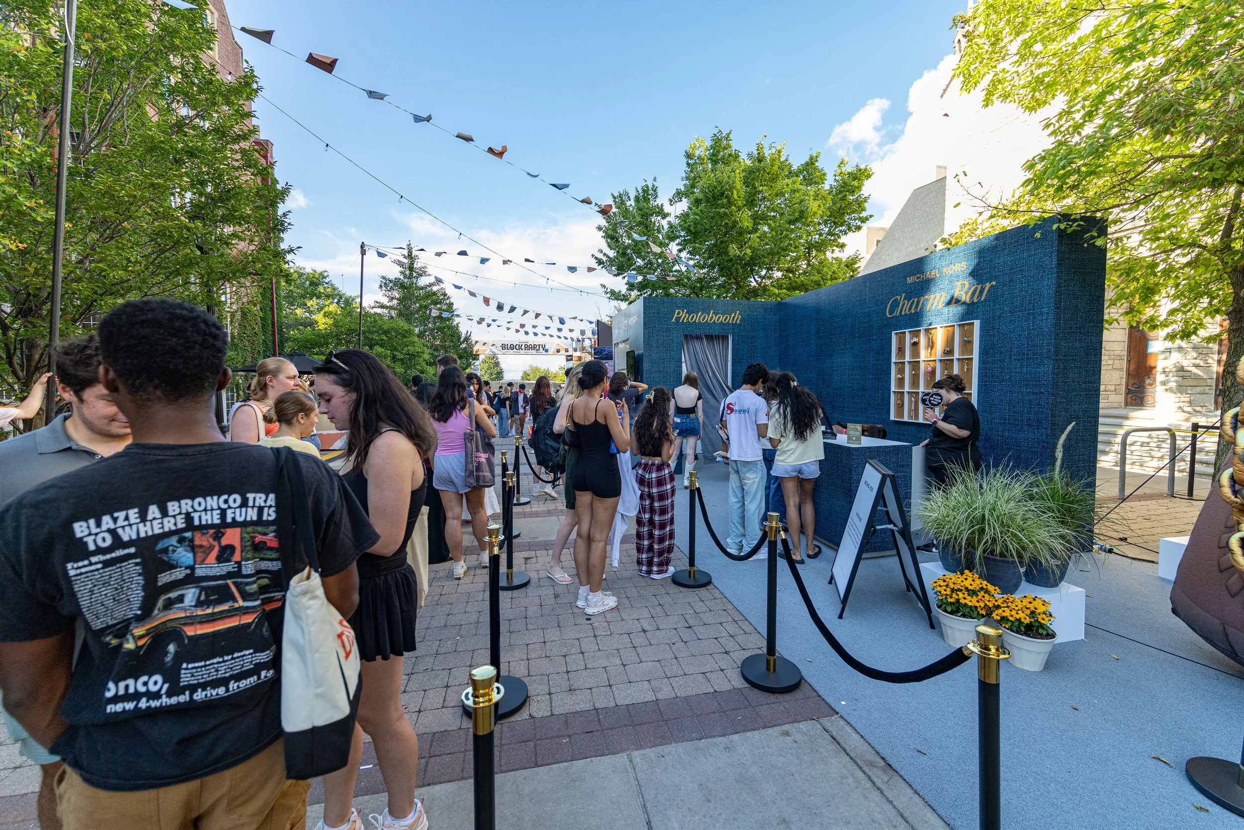 People standing in line at a photo booth and charm bar booth in an outdoor event with string flags and greenery.
