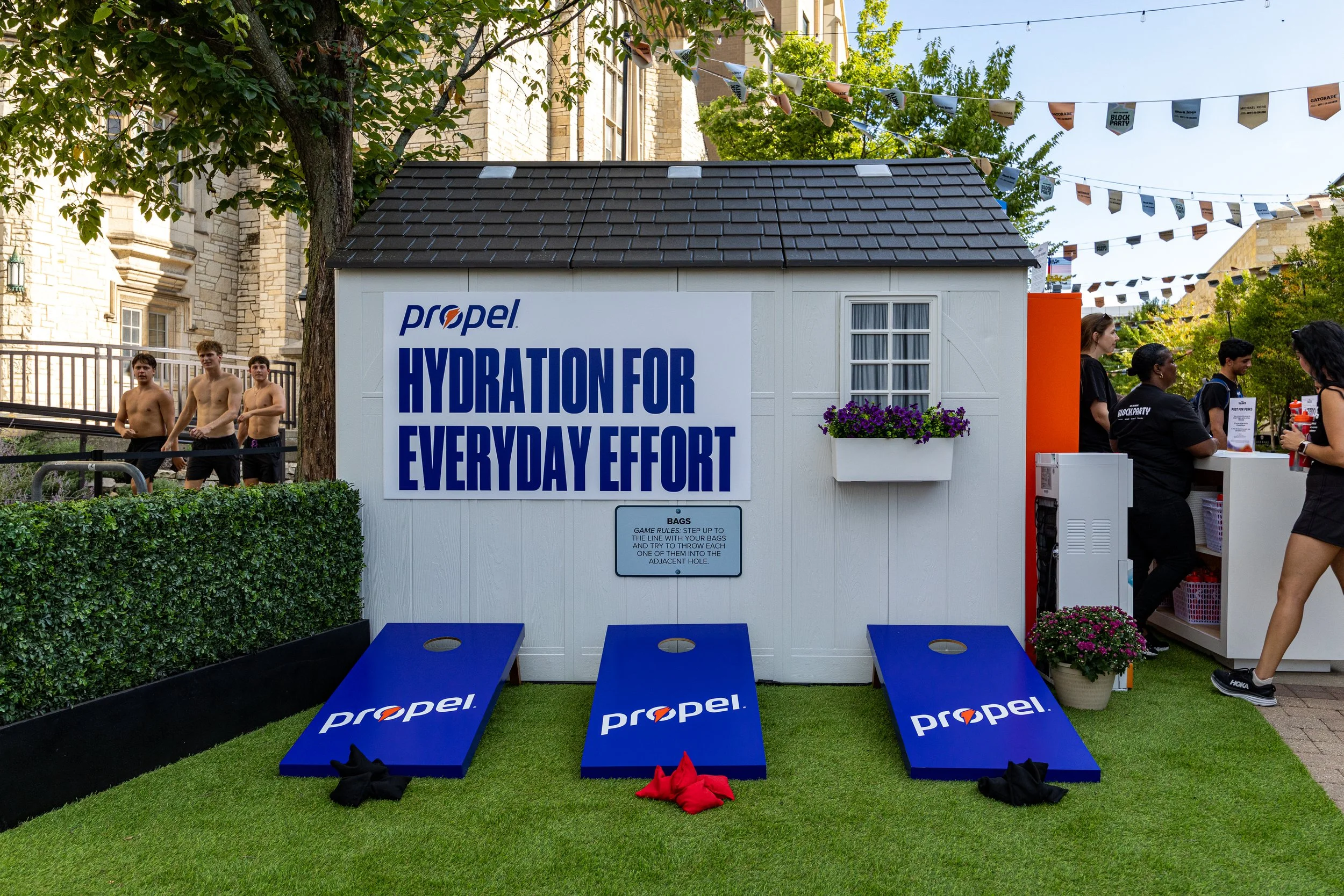 Outdoor scene of a hydration station with three blue cornhole boards featuring the Propel logo, a white shed with a large Propel sign reading 'Hydration for Every Day Effort,' and people around it. There are sprigs of purple flowers in a white plante