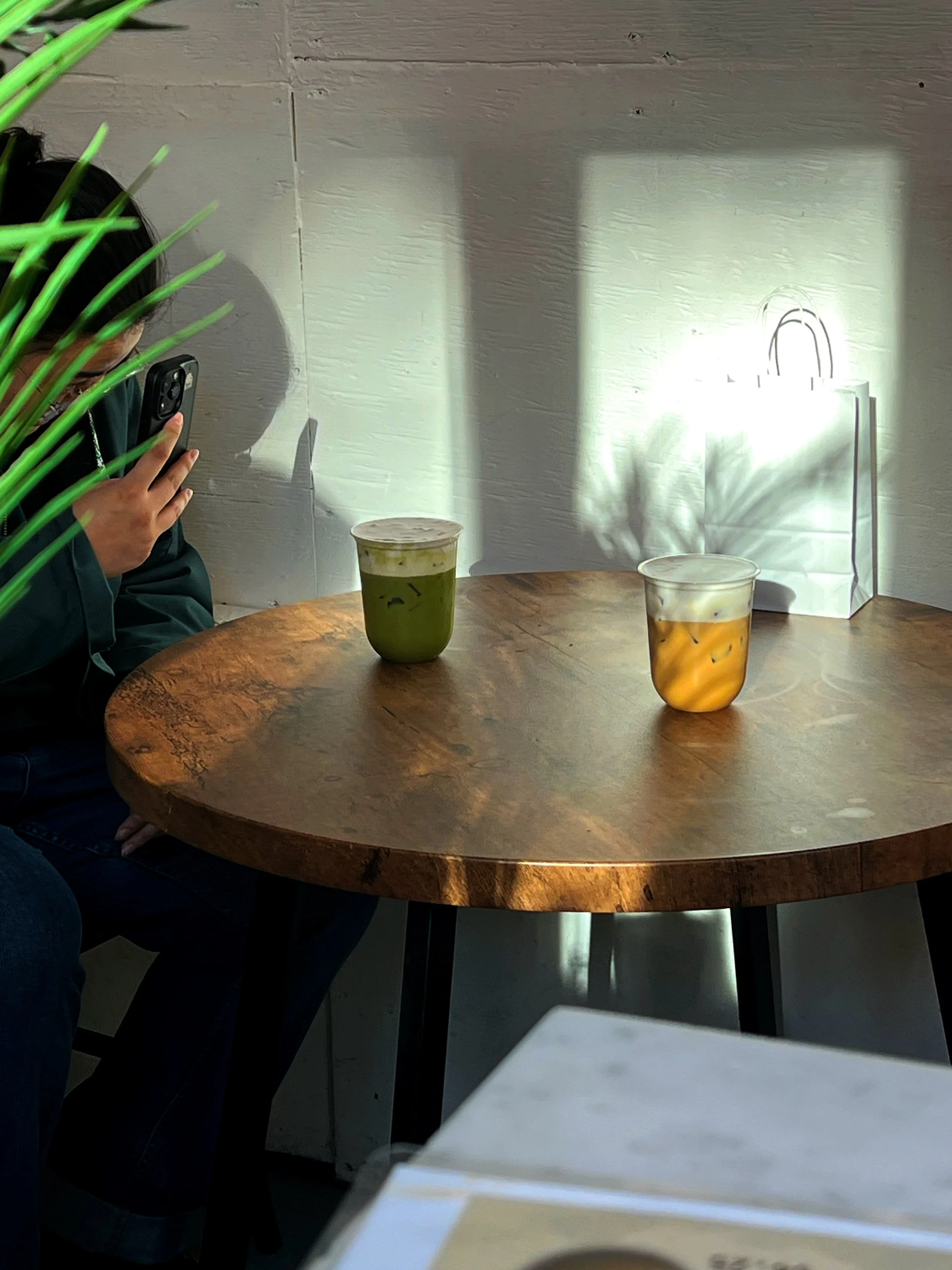 Person holding a smartphone, sitting at a round wooden table with two drinks, sunlight casting shadows on the white wall behind.Aesthetic matcha and latte picture